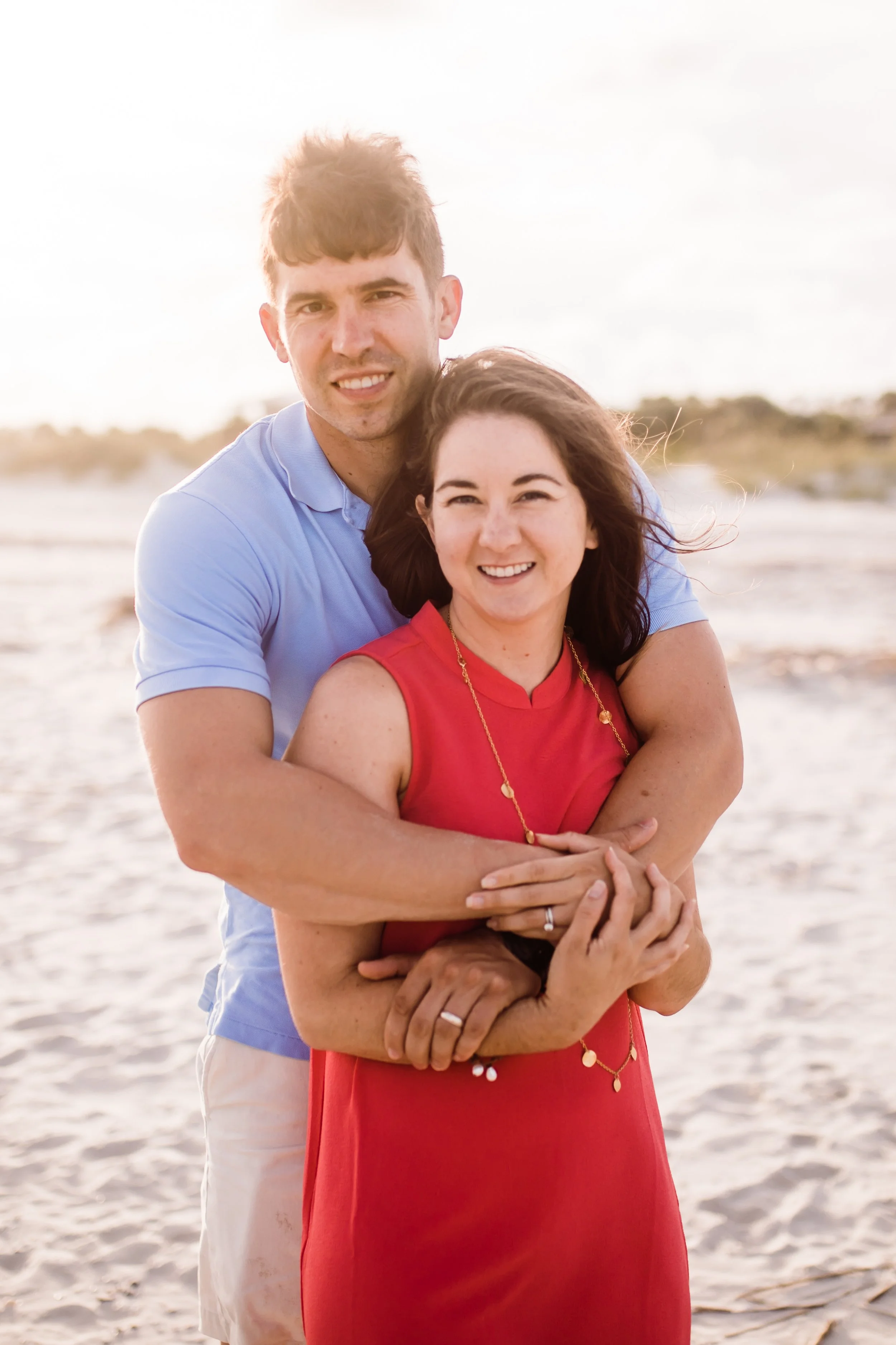 A smiling man and woman embracing on a sandy beach during sunset, with the man standing behind the woman and both looking at the camera.