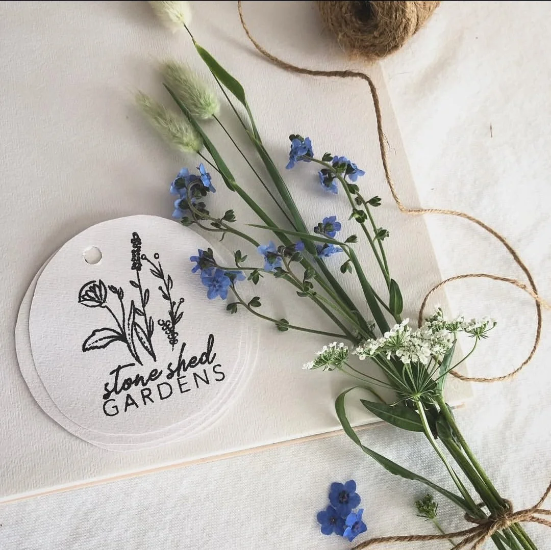A bouquet of blue and white flowers tied with twine, placed near a round white tag with the logo and words "Stone Shed Gardens" alongside black line art of flowers, on a light surface.