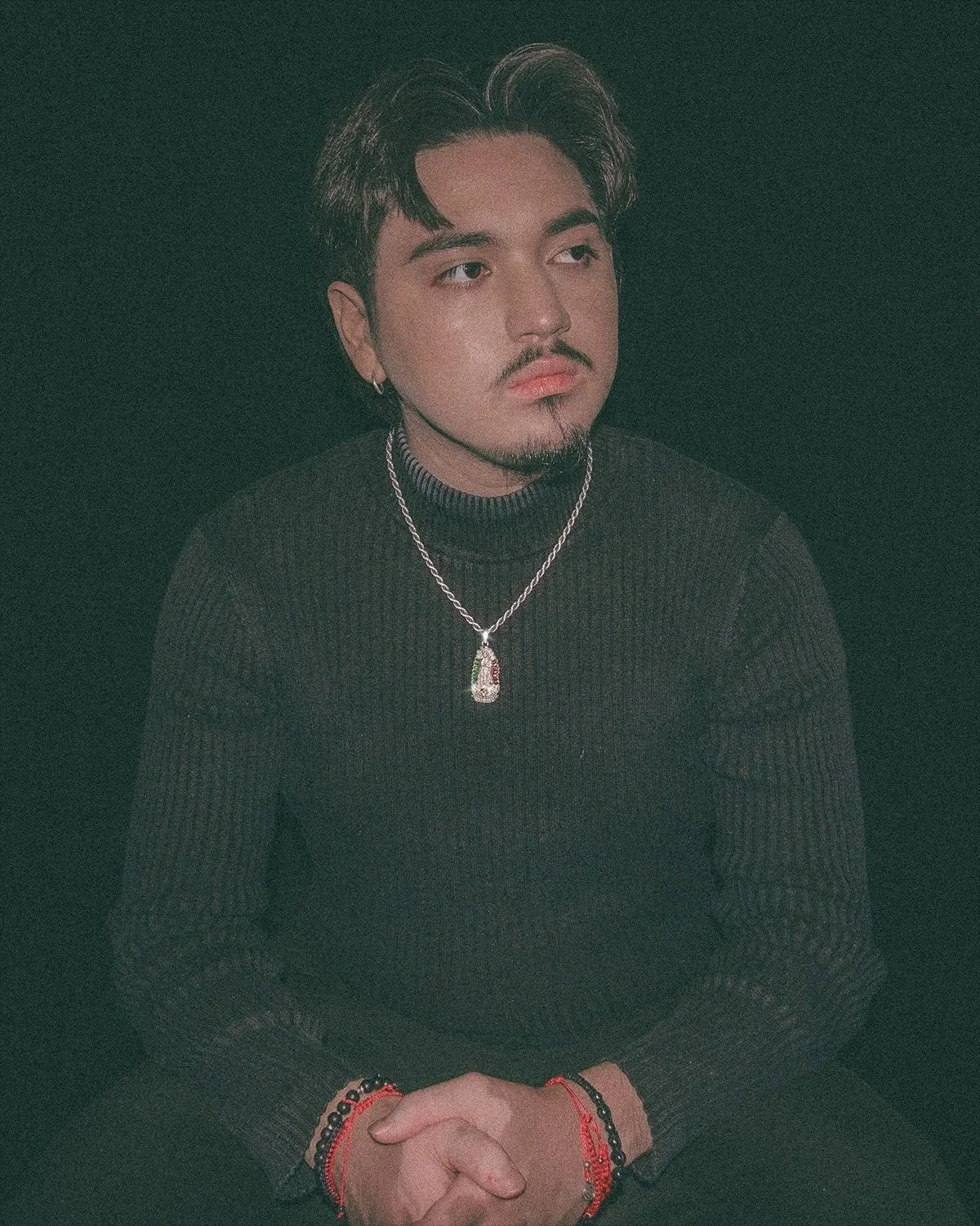 Young man with dark hair, beard, and mustache, wearing a black turtleneck sweater, silver chain with pendant, and red and black bracelets, sitting against a dark background.