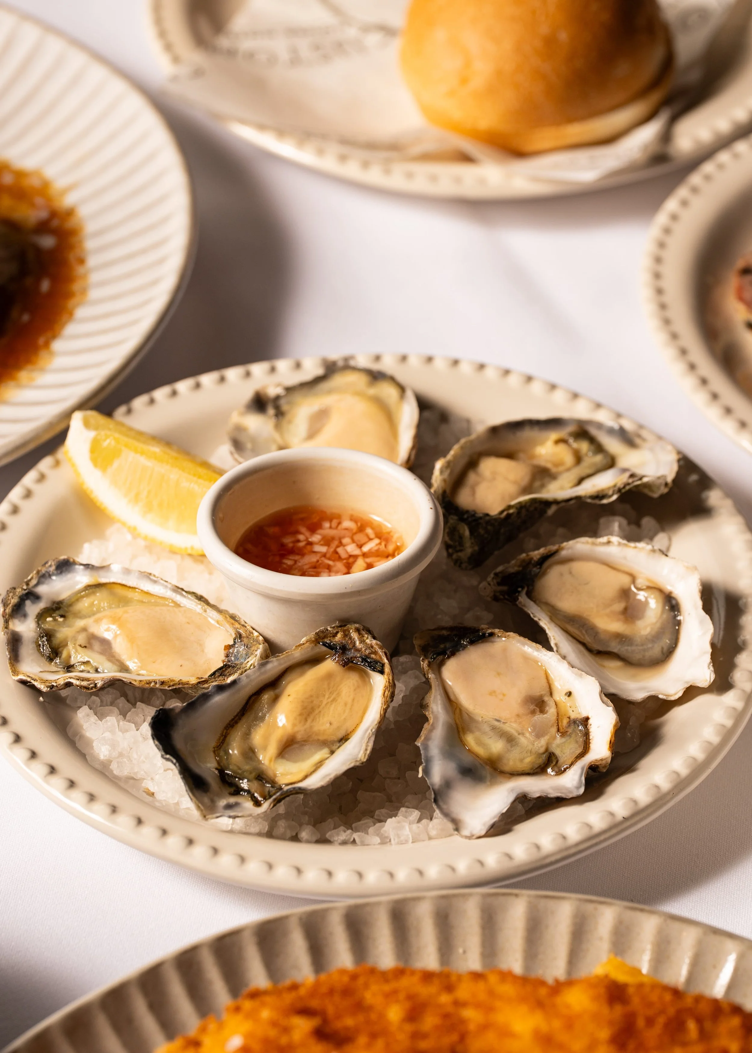 Person holding a plate of Spanish tapas with slices of cured ham, a dollop of mayonnaise or aioli, and crispy bread crisps, surrounded by other dishes on a wooden table.