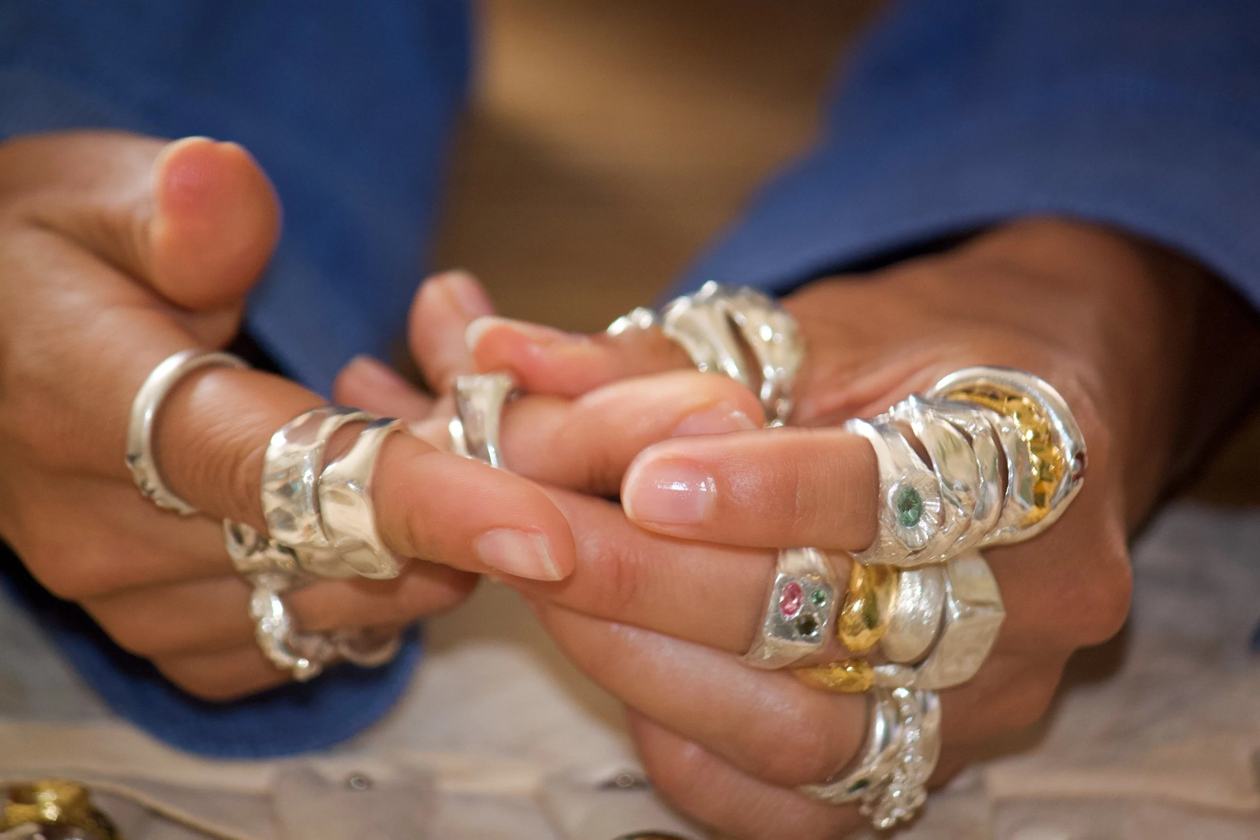 Close-up of a person's hands adorned with numerous chunky silver and gold rings, with some rings featuring small colored gemstones, against a blurred background.