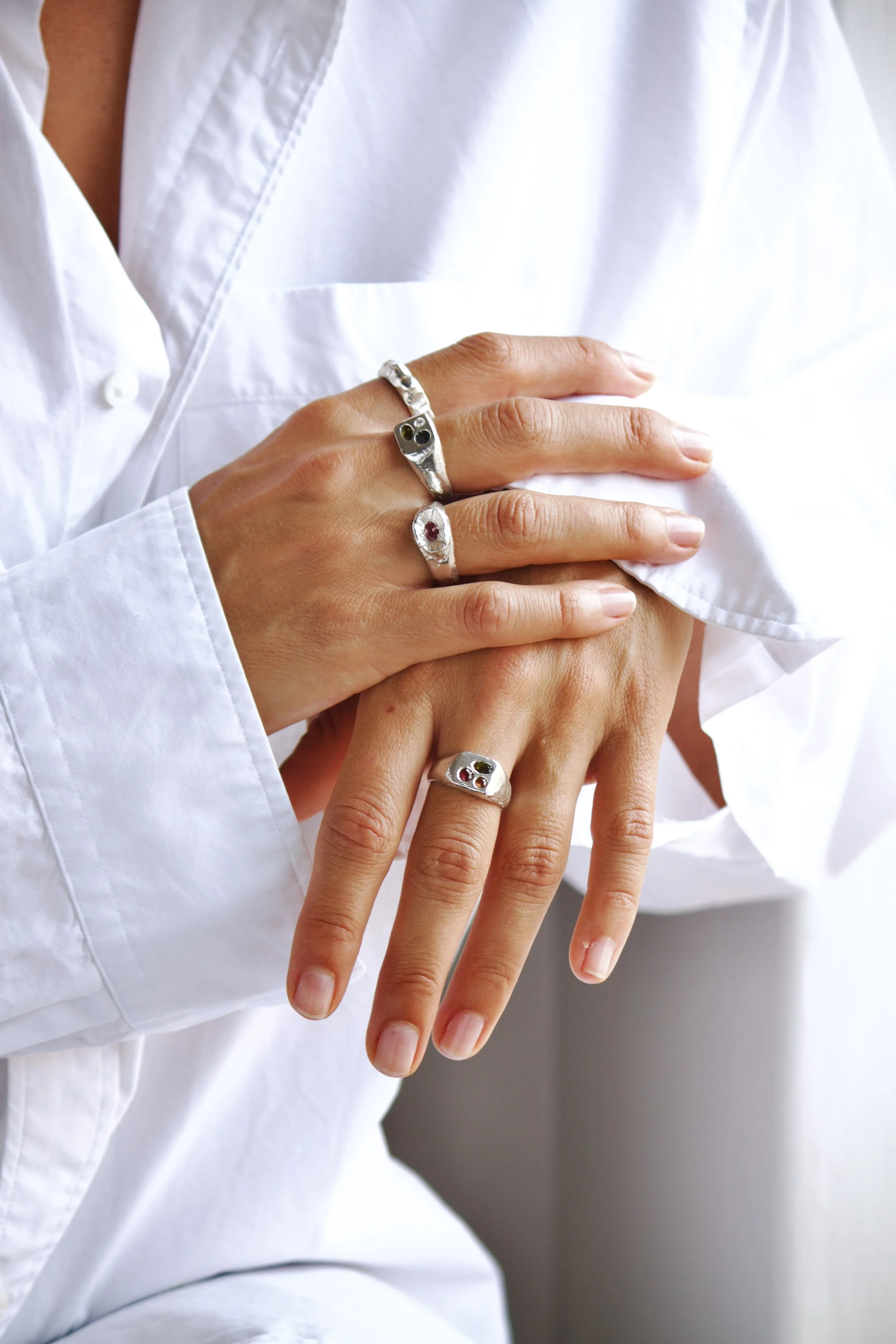 Hands wearing silver rings with colorful stones, resting on a white shirt.
