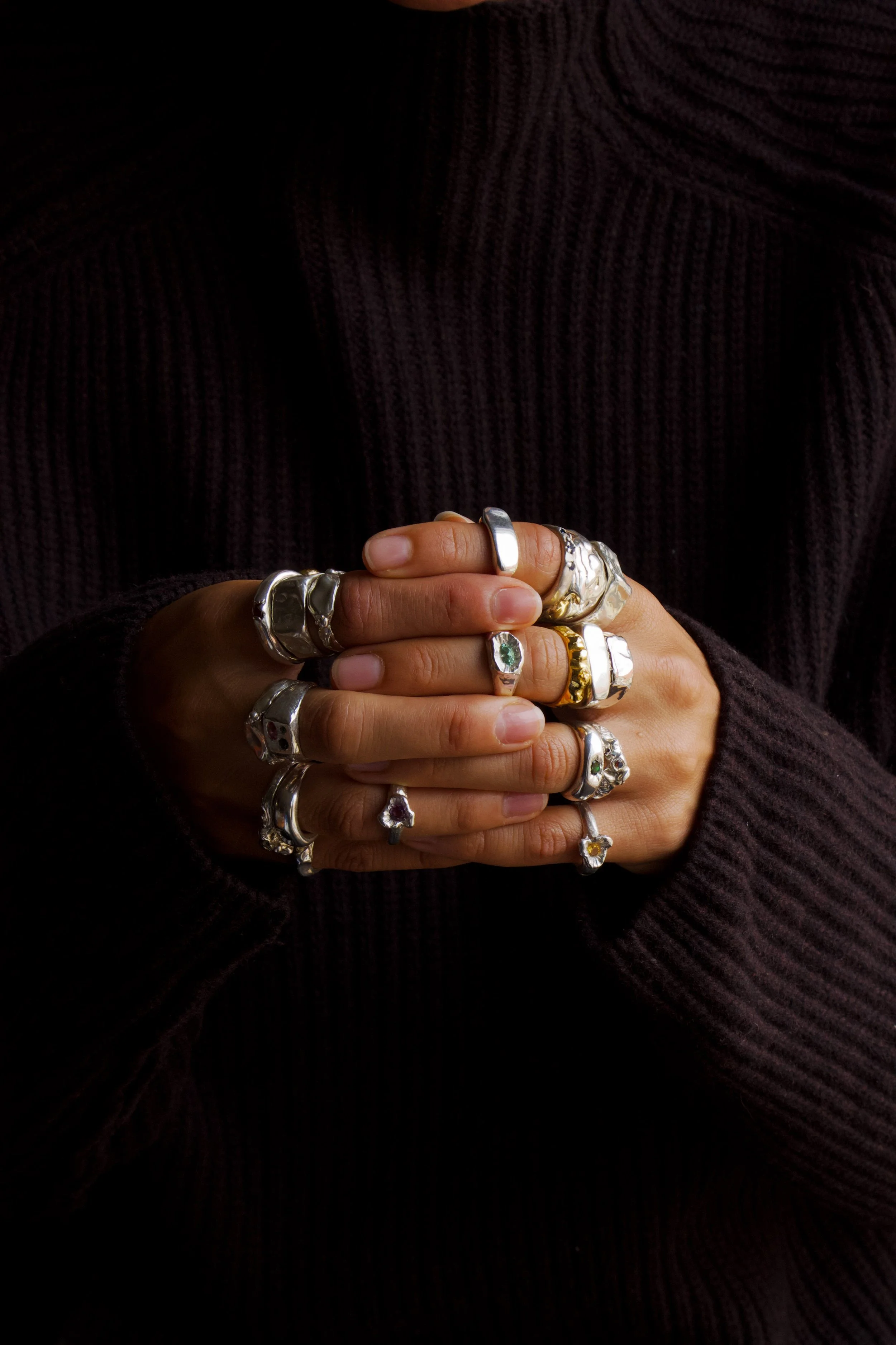 Person wearing multiple silver and gold rings with various designs and gemstones on fingers, dressed in a dark ribbed sweater, hands clasped together.