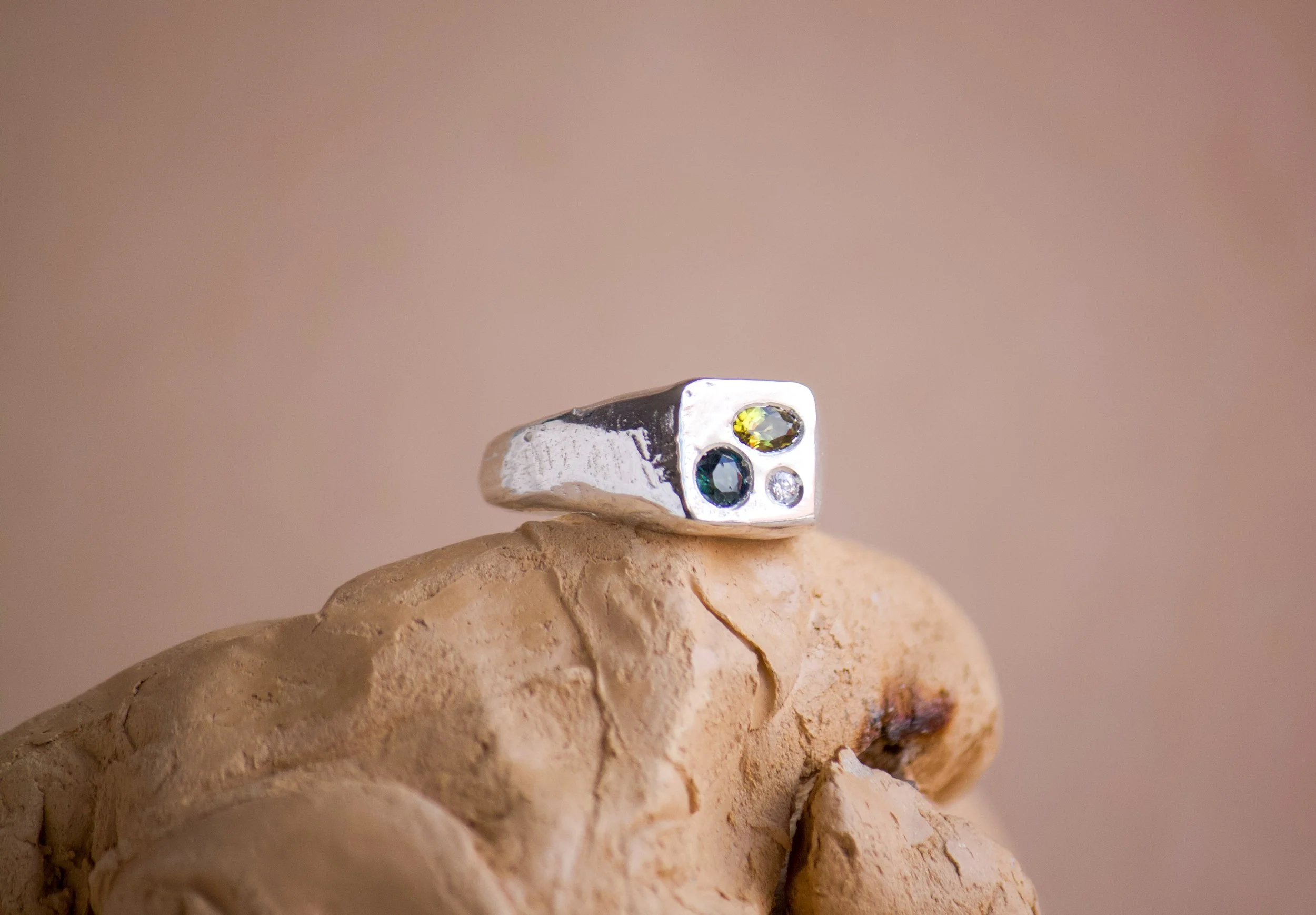Silver ring with three gemstones: a small clear stone, a larger blue stone, and a dark greenish stone, set against a plain white background.