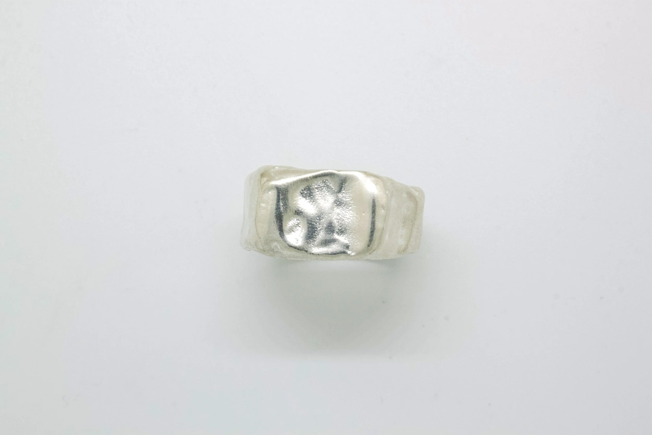 A silver metallic ring with a raised design on top, placed on a white surface.