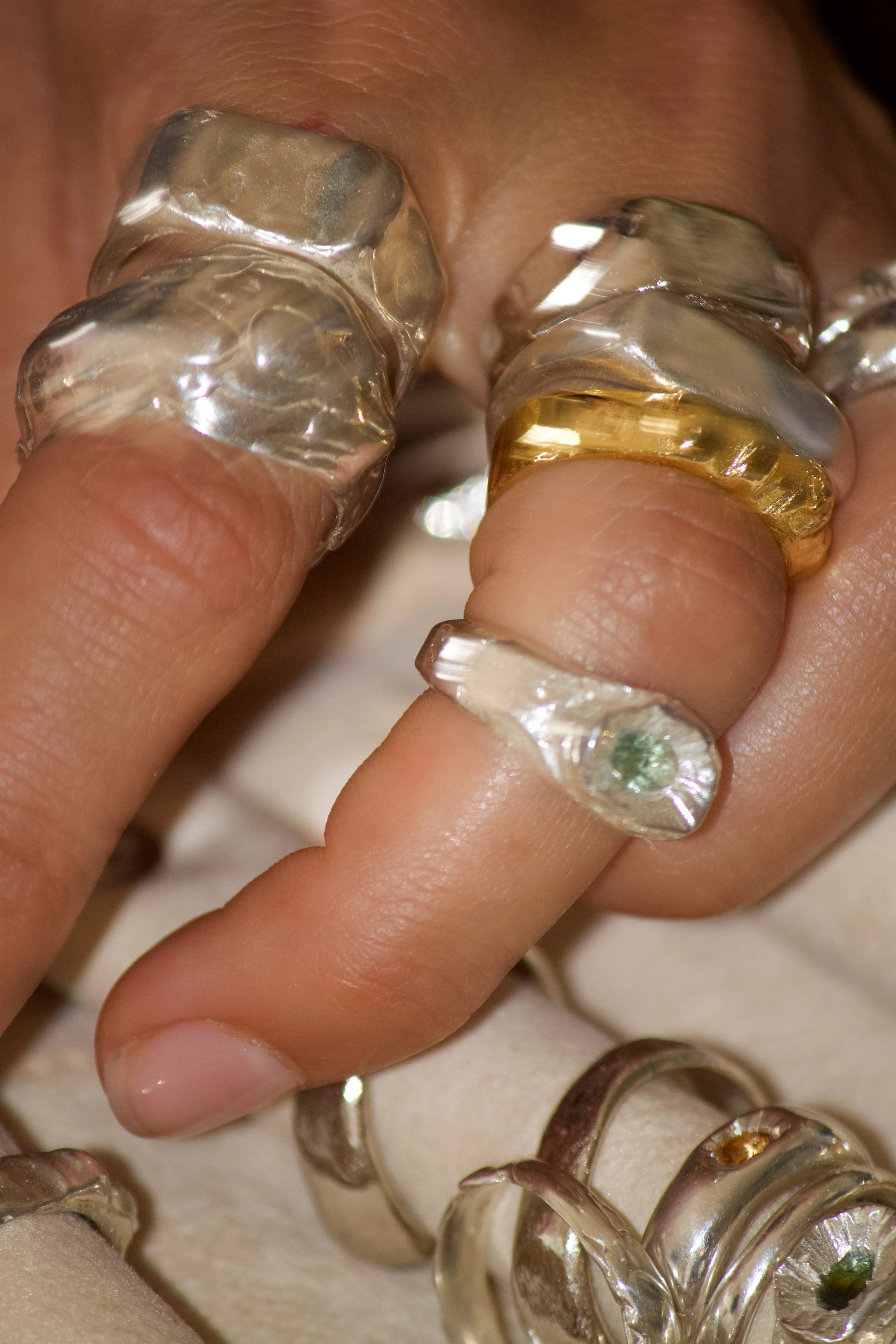 Close-up of fingers wearing assorted transparent and colored rings, with a focus on the finger with a gold-colored ring, against a blurred background.