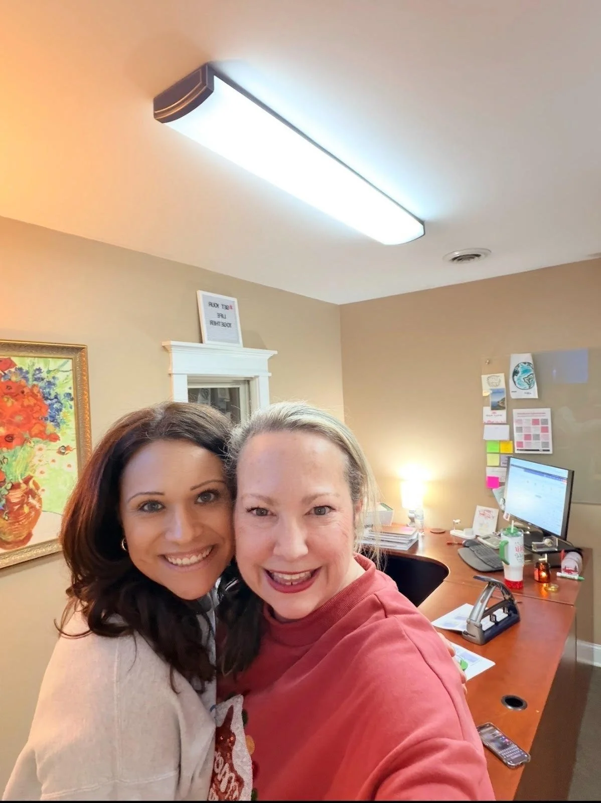 Two women smiling and taking a selfie in an office with a painting, a desk with a computer, and various office supplies and decorations in the background.