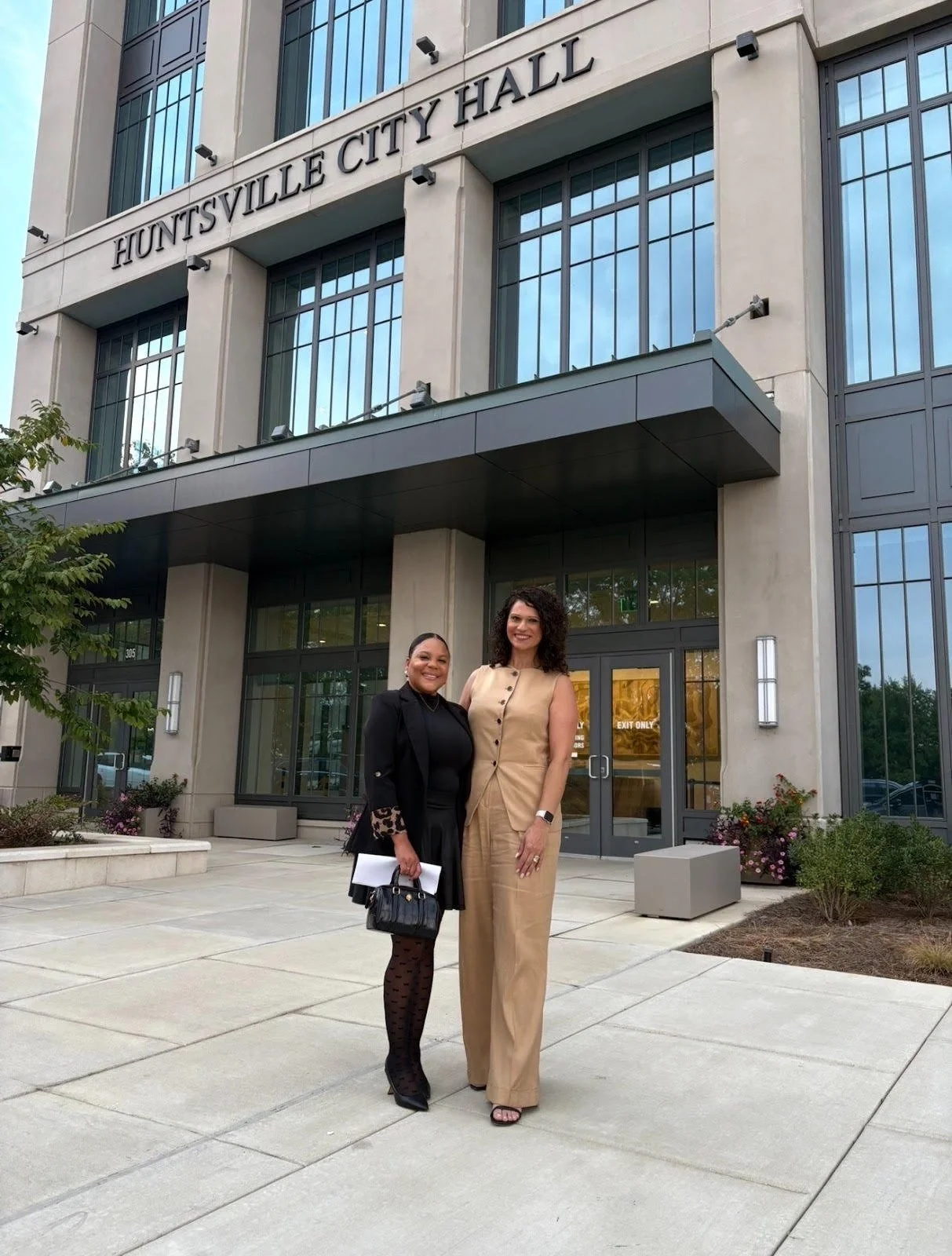 Two women standing outside Huntsville City Hall in front of the building's entrance, smiling at the camera.