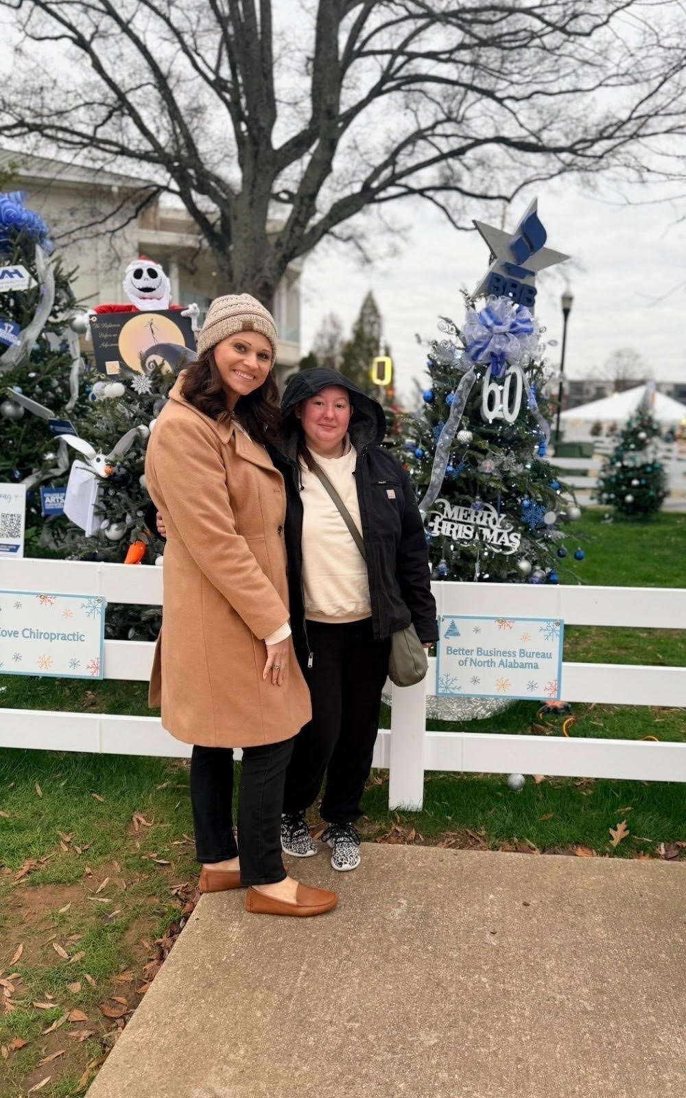 Two women standing outdoors in front of decorated Christmas trees, with signs reading 'Better Business Bureau of North Alabama' and 'Merry Christmas'. One woman is wearing a tan coat and beige knit hat, the other is in a black jacket with a hood. They are smiling at the camera on a cloudy day.