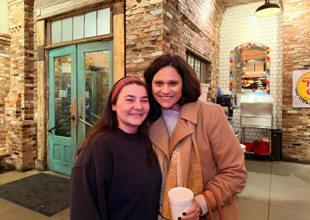 Two women smiling inside a restaurant with exposed brick walls and a colorful mirror in the background. One woman has long brown hair and wears a black shirt, and the other has shoulder-length dark hair, a beige jacket, and holds a white cup.