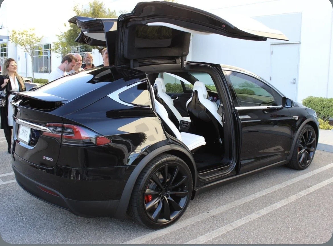 Black electric car with gull-wing doors open, showing white interior seats, parked outdoors with a group of people nearby.
