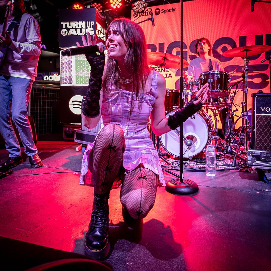 Lyric performing on stage, kneeling in front, holding a microphone, wearing black fishnet stockings, knee-high lace-up boots, and black gloves, with a drum set and large screen in the background.