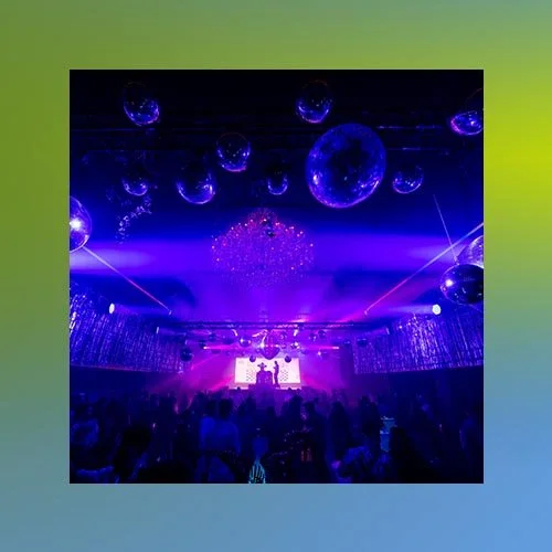 A nightclub or concert venue with purple and blue lighting, large disco balls hanging from the ceiling, and a crowd of people facing a stage with a DJ or performer.
