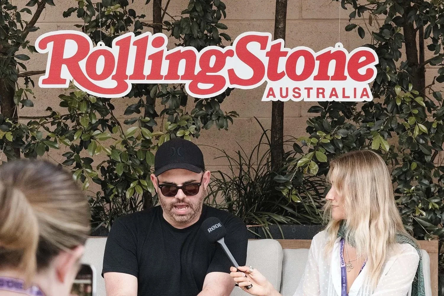 A man being interviewed by a woman at the Rolling Stone Australia event, with the Rolling Stone Australia sign and greenery in the background.