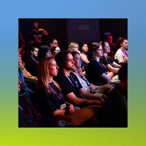 Audience members sitting and listening attentively at a conference with dark lighting and colorful accents.