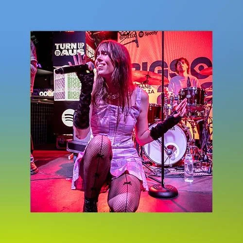 A female singer performing on stage with a band, holding a microphone, wearing fishnet stockings, black gloves, and a dress, with a drummer and background music screens.