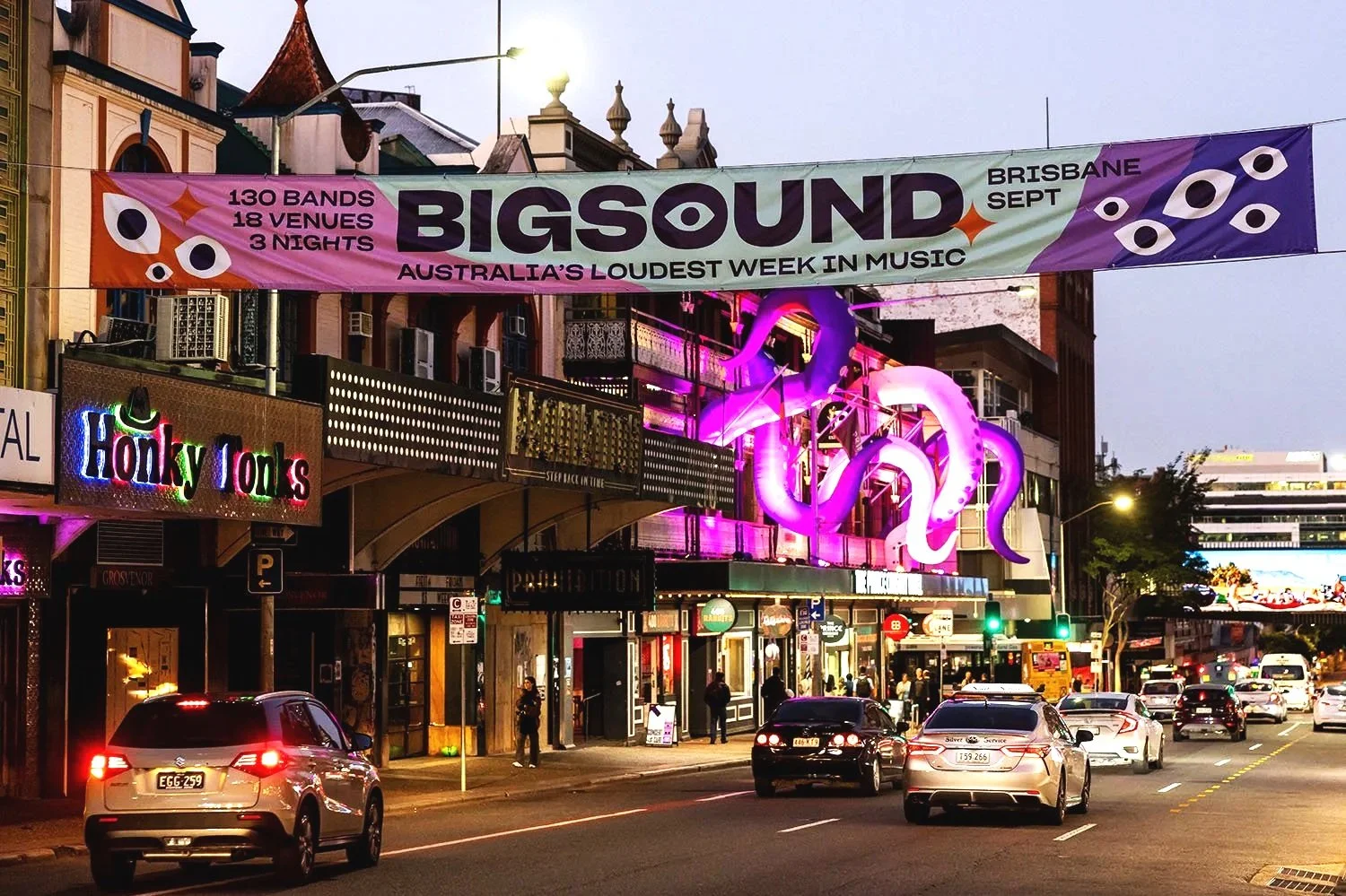 City street with neon signs, a large colorful banner for BIGSOUND music festival, illuminated pink octopus sculpture on building, cars on road, pedestrians walking, evening setting.