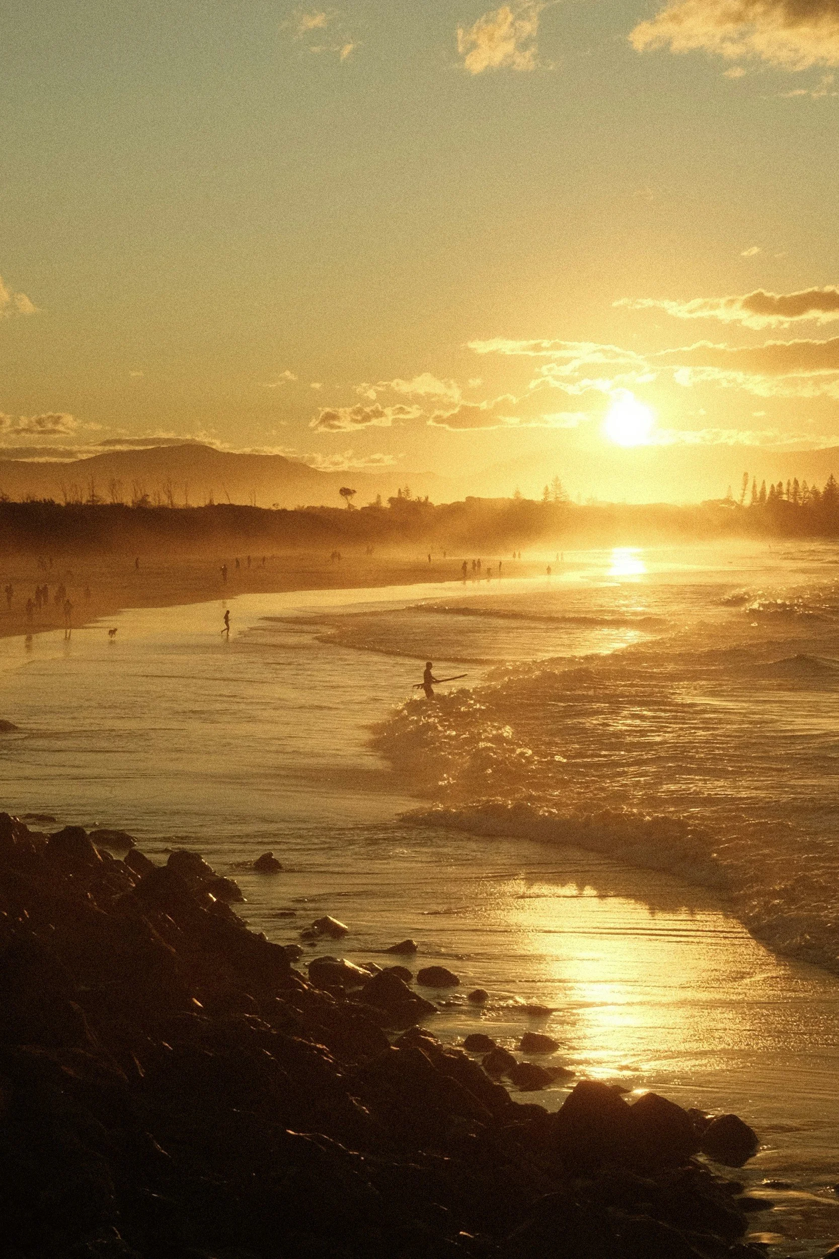 Sunset over a beach with people walking and surfing, rocks in the foreground, and a golden sky.