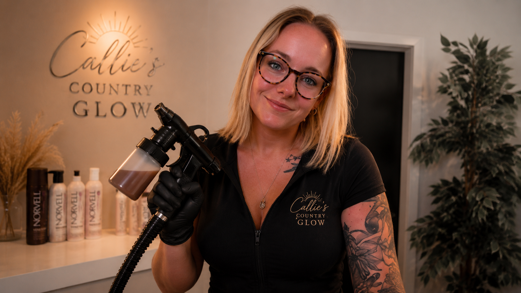 A woman with blonde hair, glasses, and tattoos on her arm holding a spray gun, smiling at the camera inside a beauty or skincare salon with a sign that reads 'Callie's Country Glow' in the background.