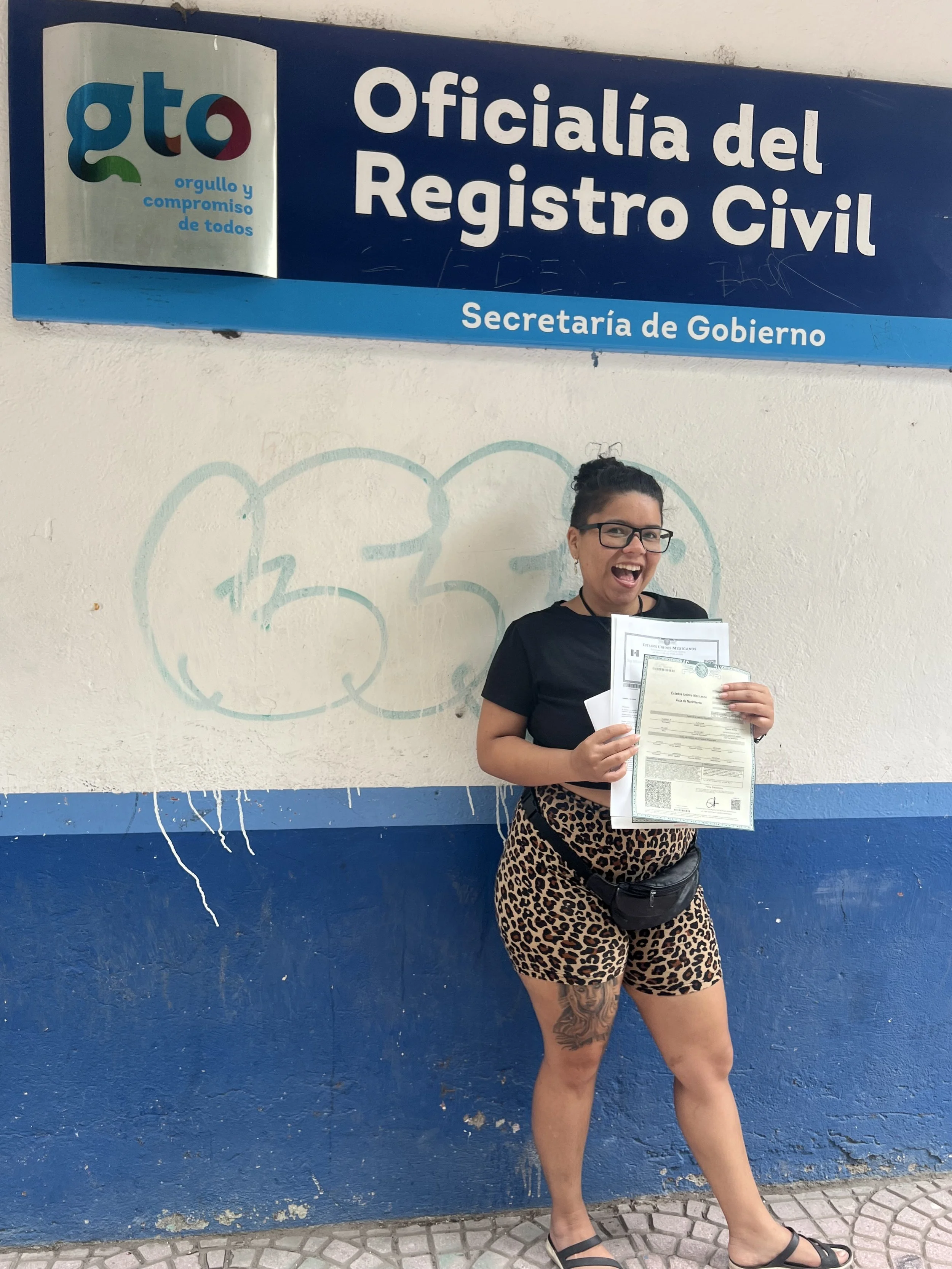 Woman standing in front of a government building with a sign that reads 'Oficialía del Registro Civil, Secretaría de Gobierno.' She is holding multiple documents and smiling.