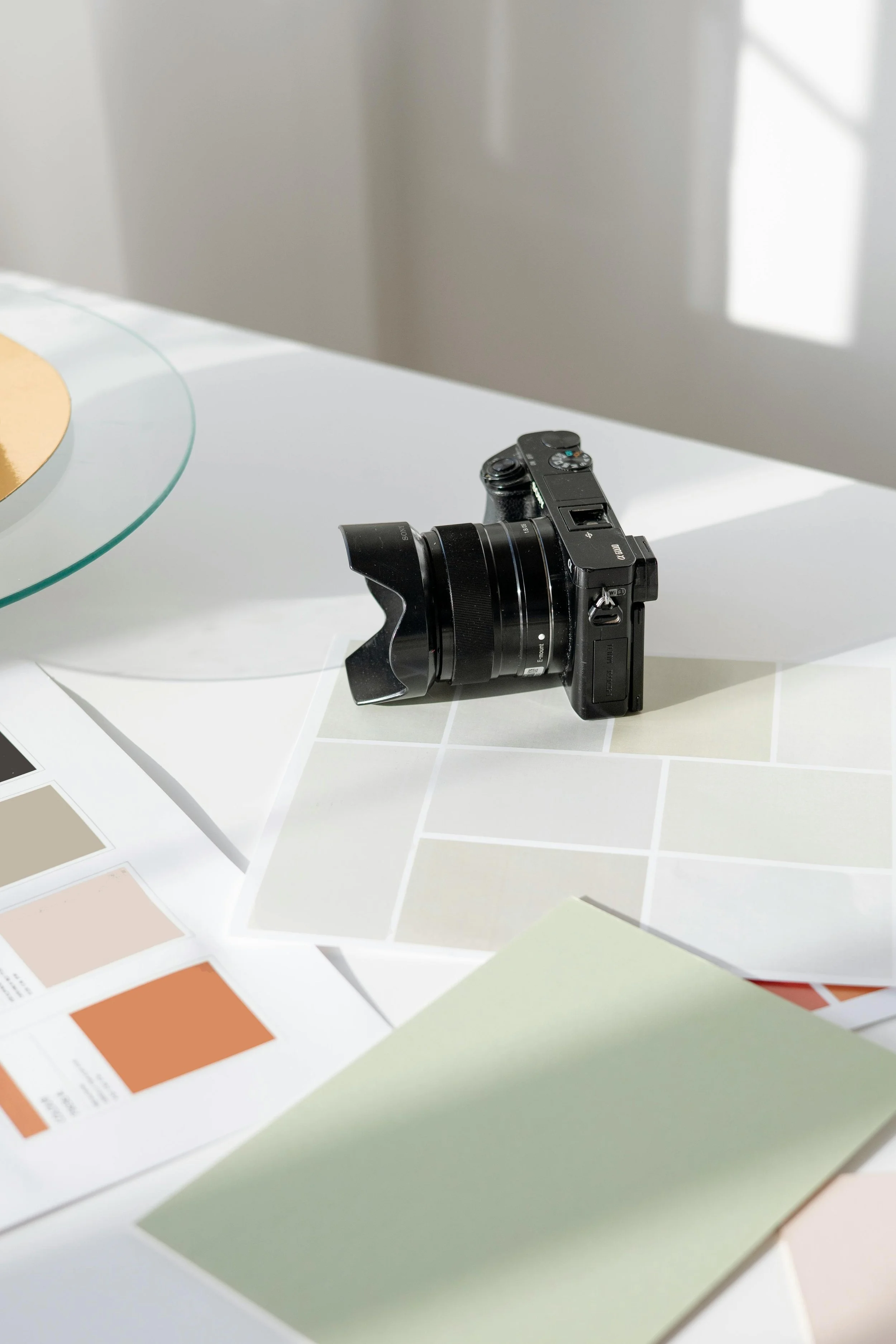 A black camera with a lens mounted on a white table surrounded by color swatches and paint samples, with sunlight casting shadows in the background.