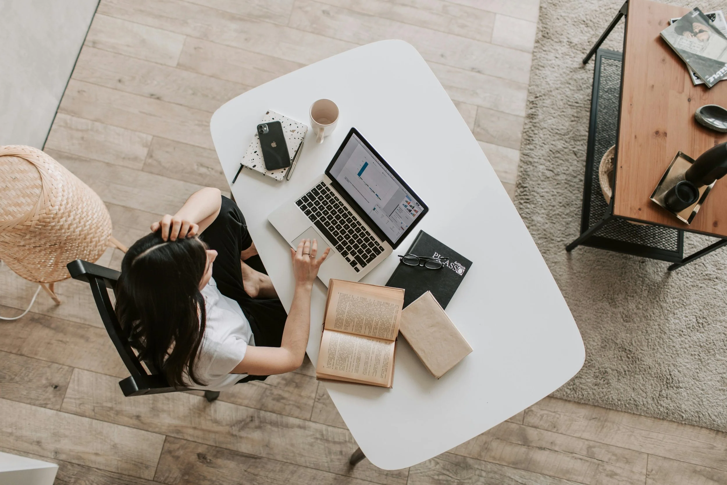 Top view of a woman sitting at a white desk working on a laptop with an open spreadsheet; the desk has a coffee mug, notebooks, a smartphone, eyeglasses, a book, and a black notebook. A wicker chair is beside the desk, and a wooden coffee table with magazines and decorative items is nearby. The room has wooden flooring and a gray area rug.