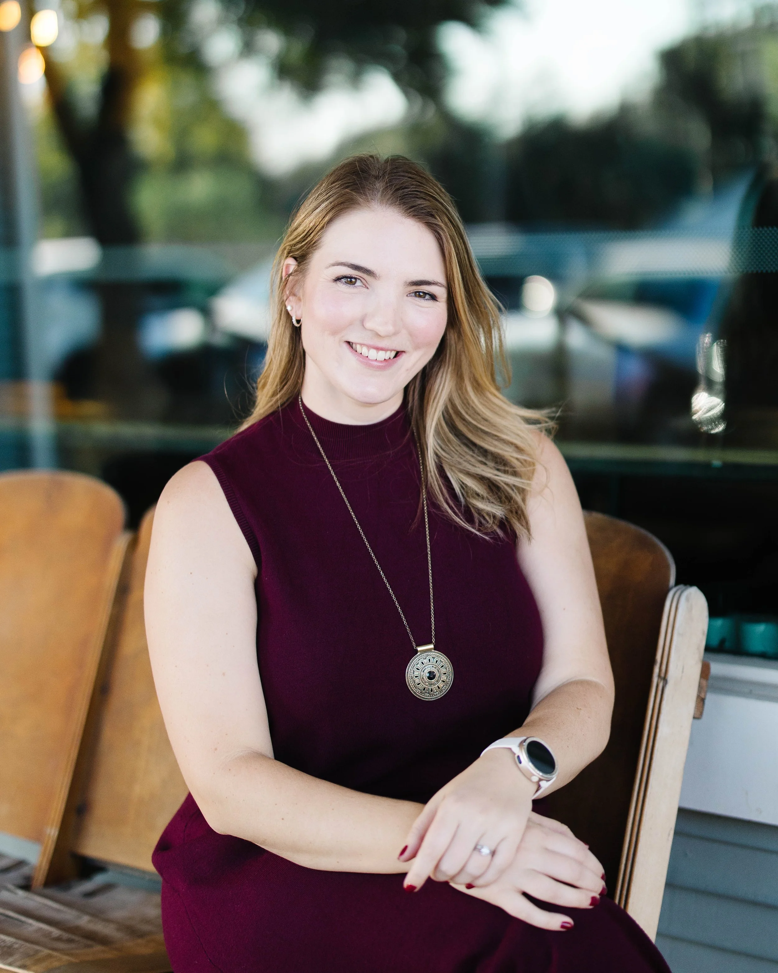 Young woman with light brown hair, smiling, wearing a sleeveless maroon top, a round pendant necklace, and a white smartwatch, sitting at a table inside a cafe with wooden chairs and a blurred background with trees outside.