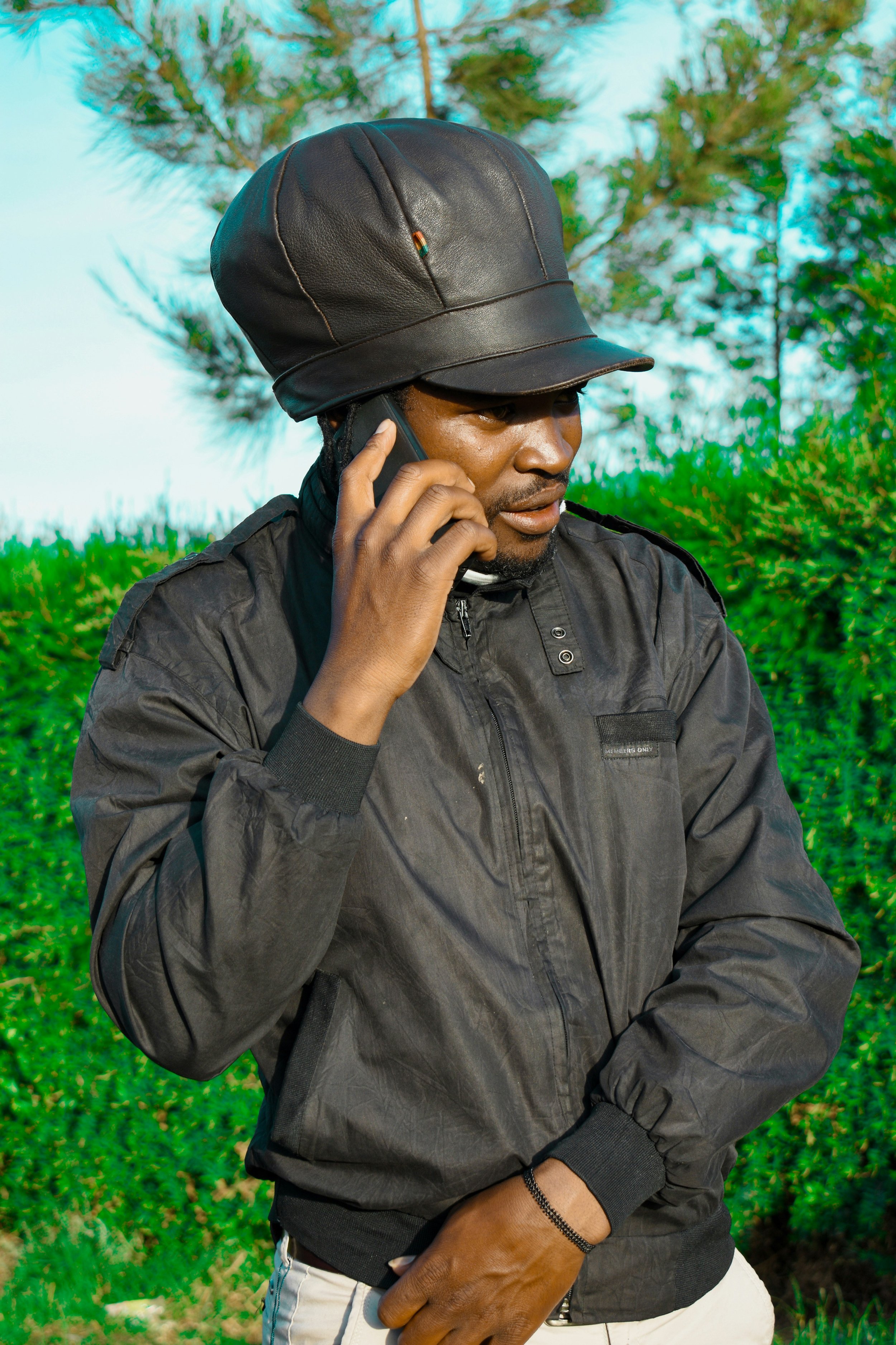 A man wearing a black leather cap and jacket is speaking on a cellphone outdoors with green trees and blue sky in the background.
