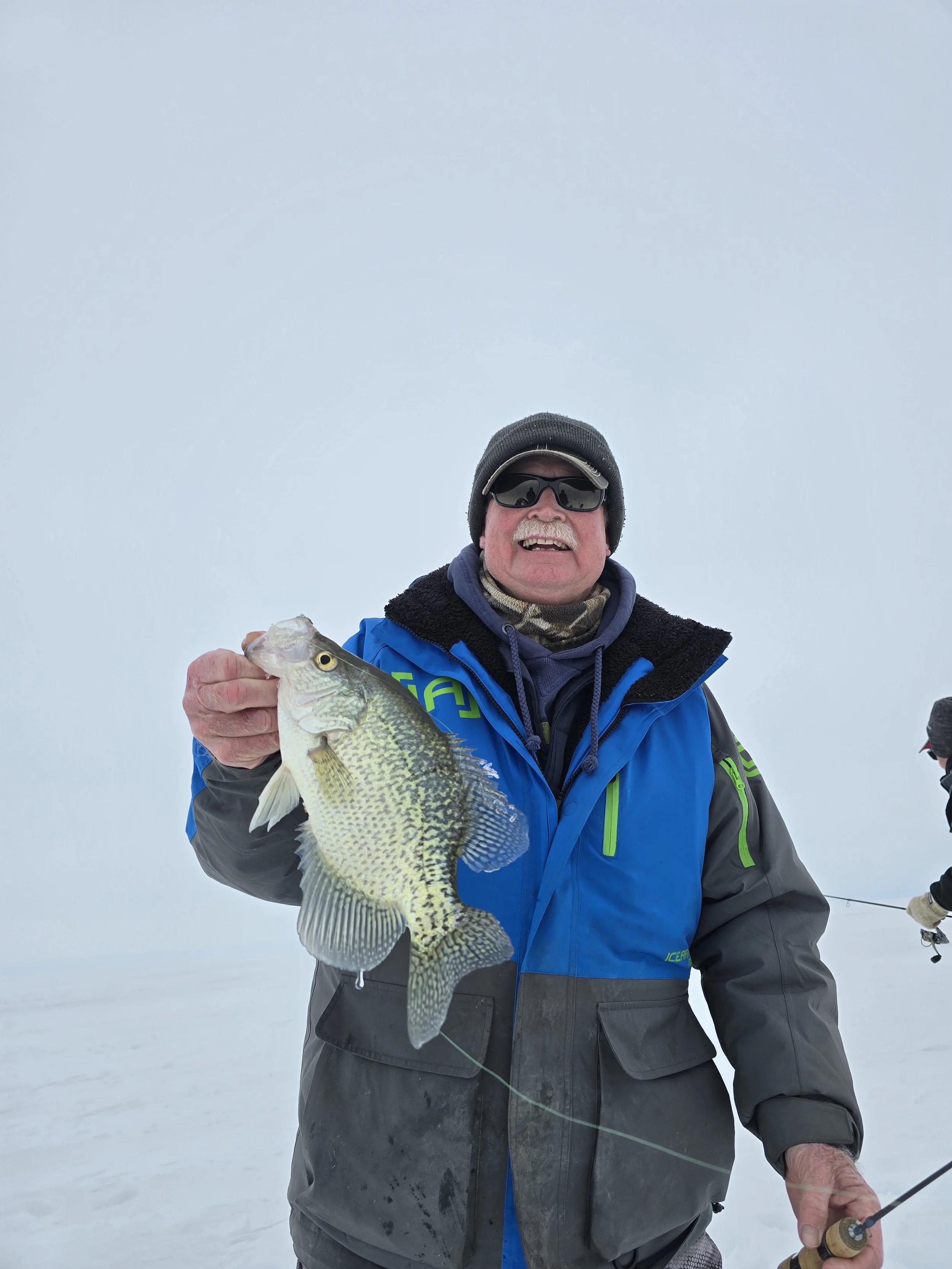 An older man in winter clothing, sunglasses, and a cap holding a large fish in one hand and a fishing rod in the other, standing on snow.