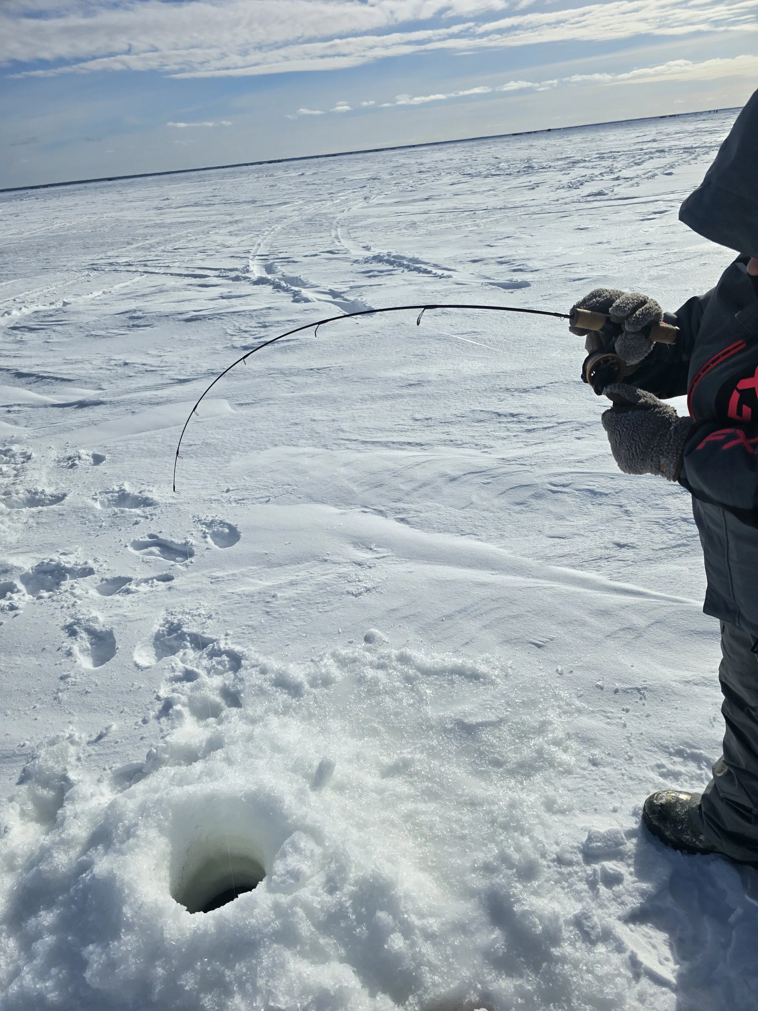 Person dressed in winter clothing sitting on snow-covered ice, holding a fishing rod, fishing through a hole in the ice on a snowy landscape with overcast sky.