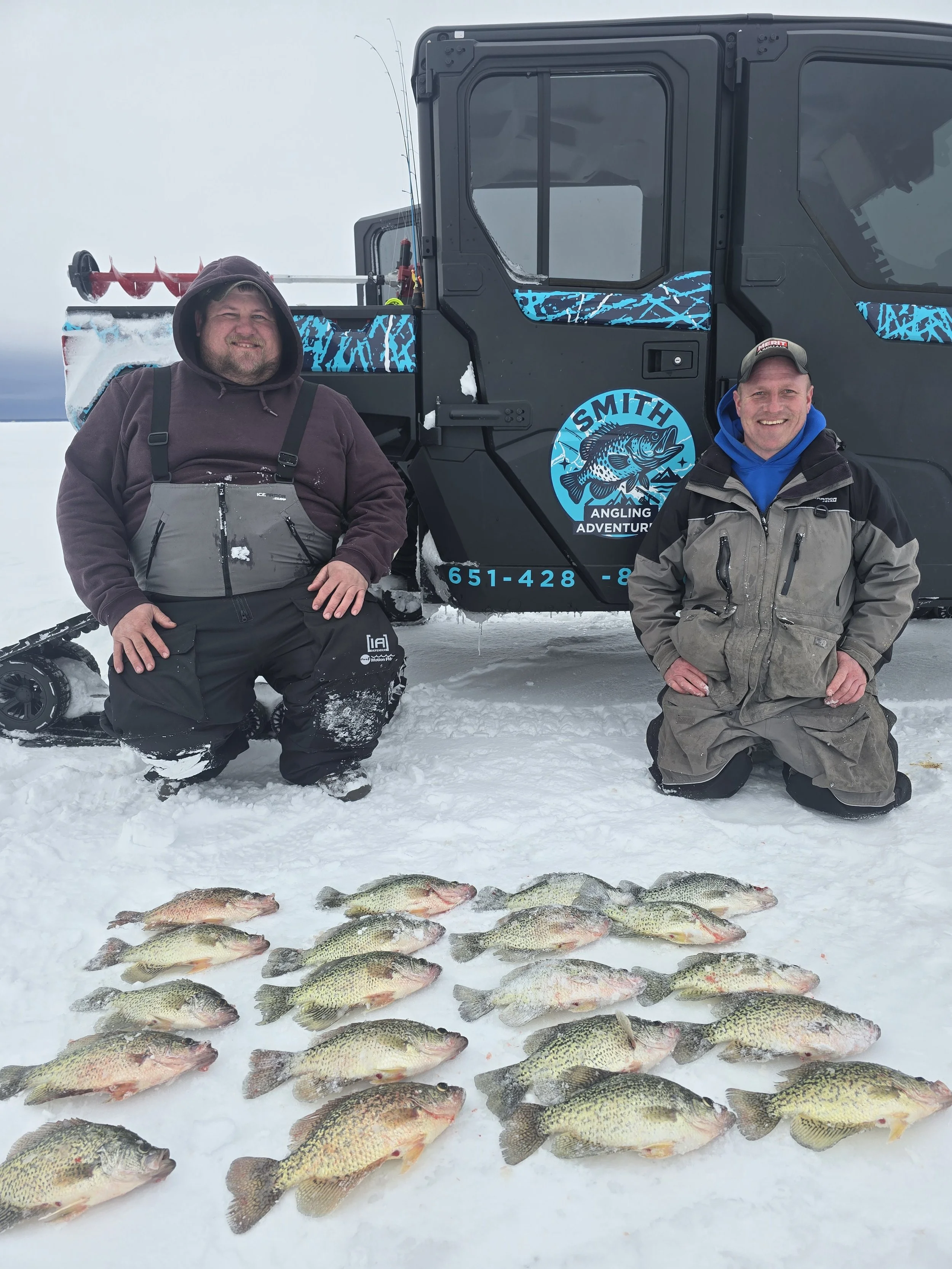 Two men kneeling in snow in front of a black snowmobile with a blue and black logo that says "Smith Angling Adventure" and a phone number. The men are dressed in winter clothing and are smiling. There are multiple fish laid out on the snow in front o