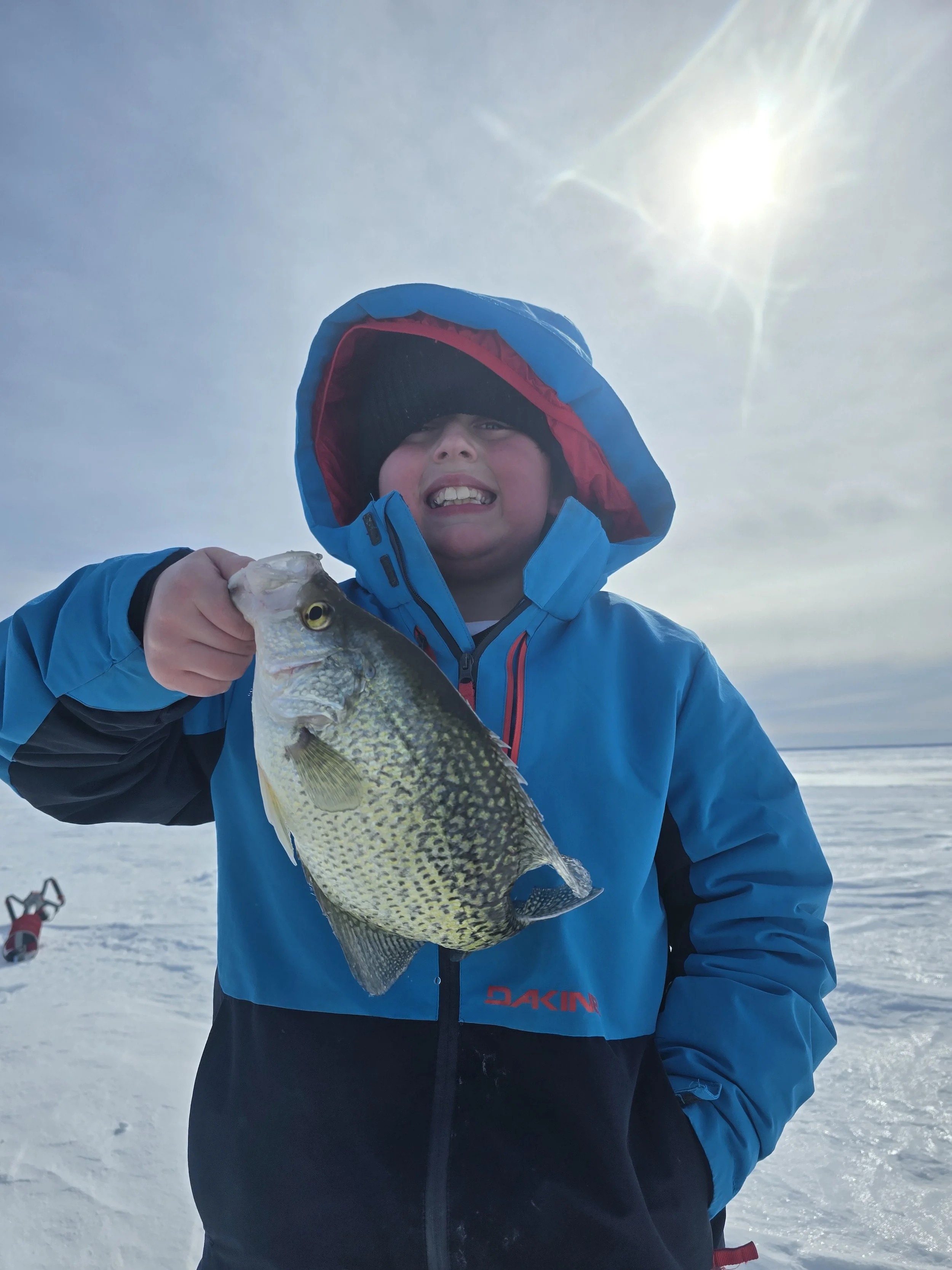 Young boy in winter jacket holding a fish, standing on snowy landscape under bright sunlight.
