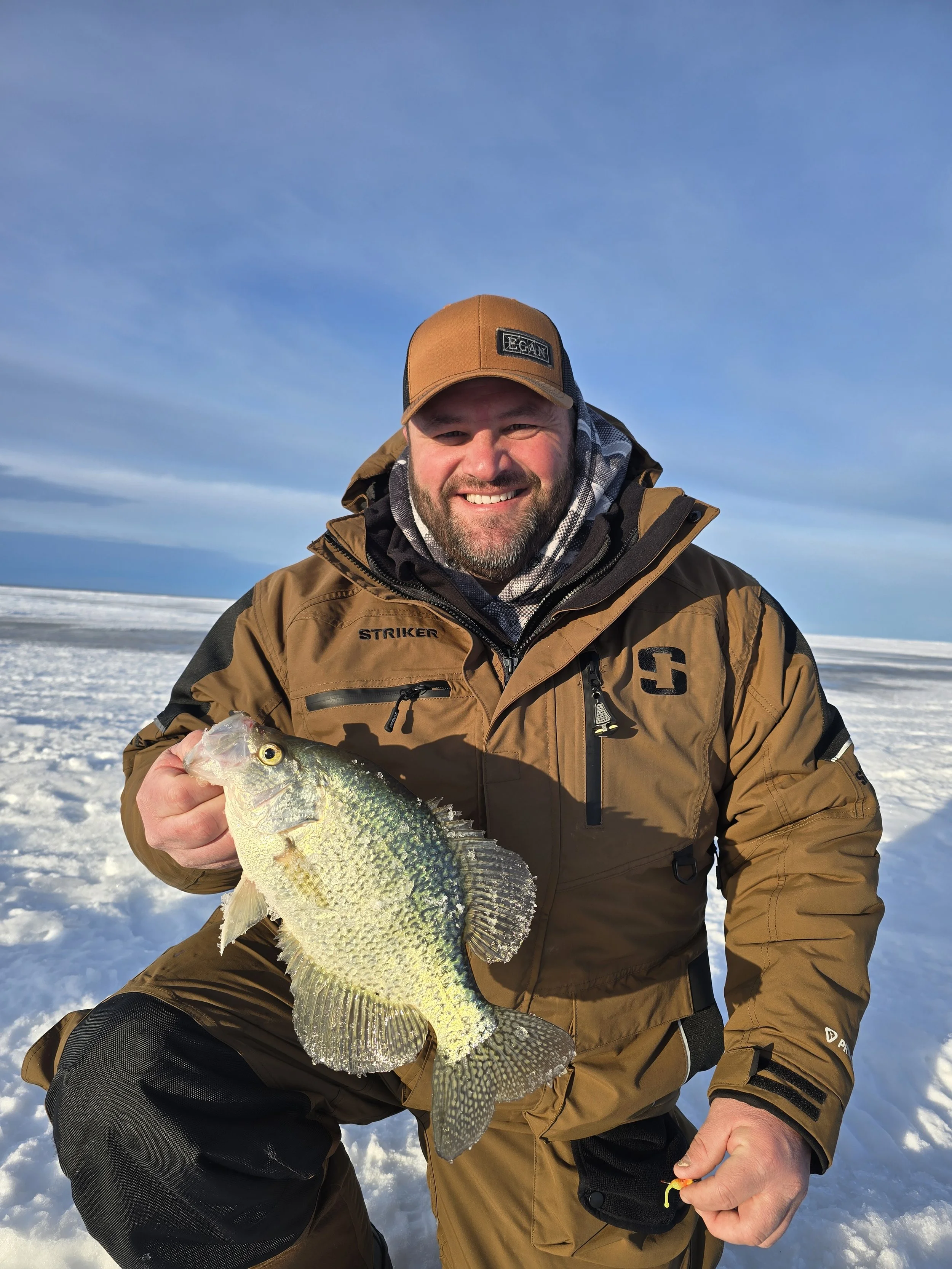 Man in brown outdoor gear kneeling on snow, holding a largemouth bass he caught while ice fishing, with a clear blue sky in the background.