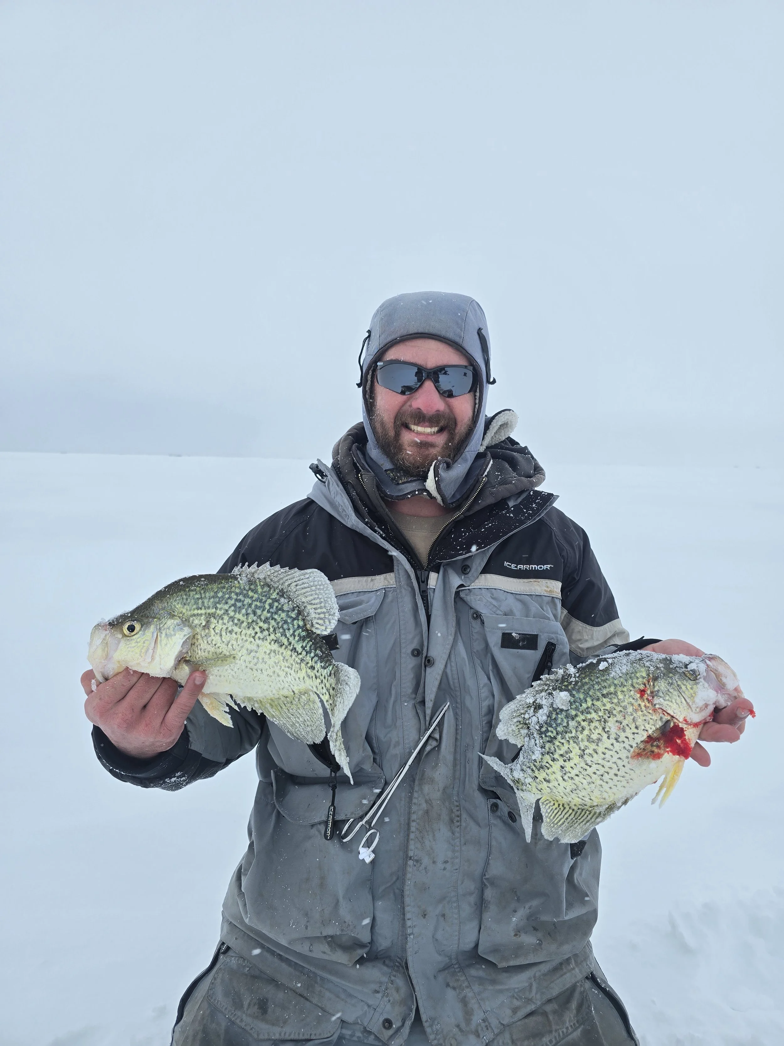 Man dressed in winter gear holding two large fish on a snowy landscape, with a cloudy sky.