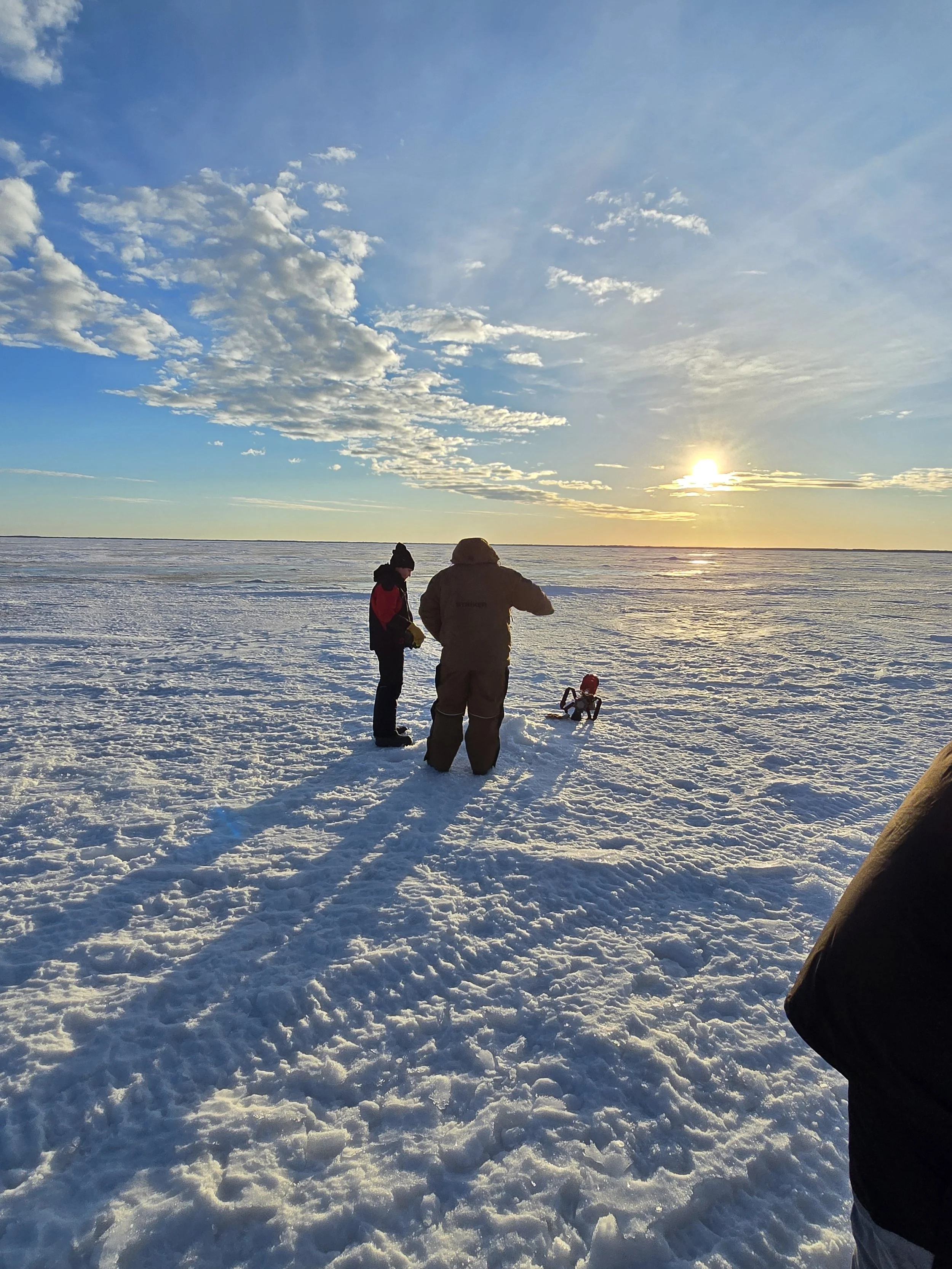Two people standing on snow-covered ground near a large body of water during sunset, with clouds in the sky.