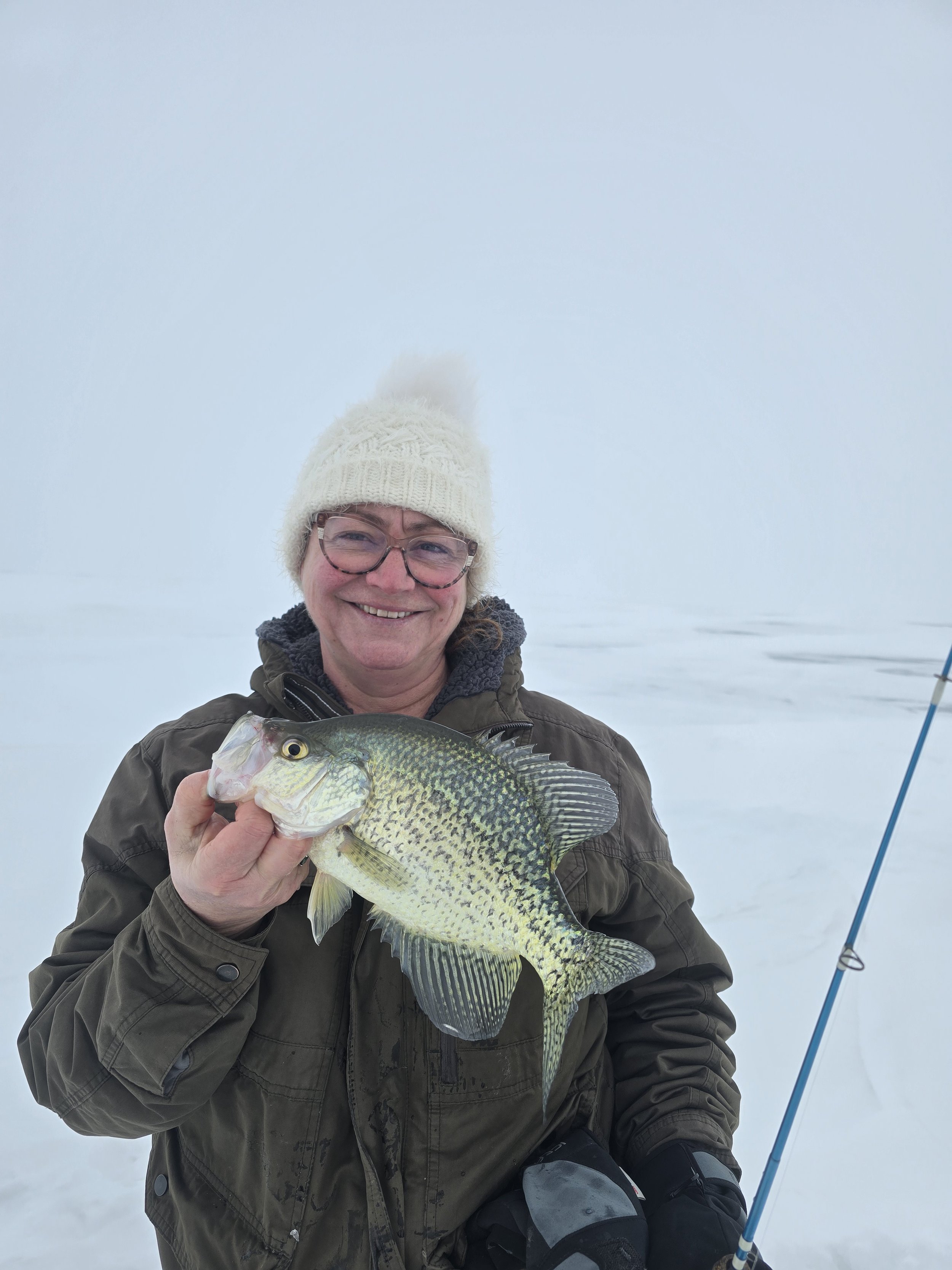 A person wearing a white knit hat and glasses holding a fish, with a snowy landscape and fishing rod in the background.