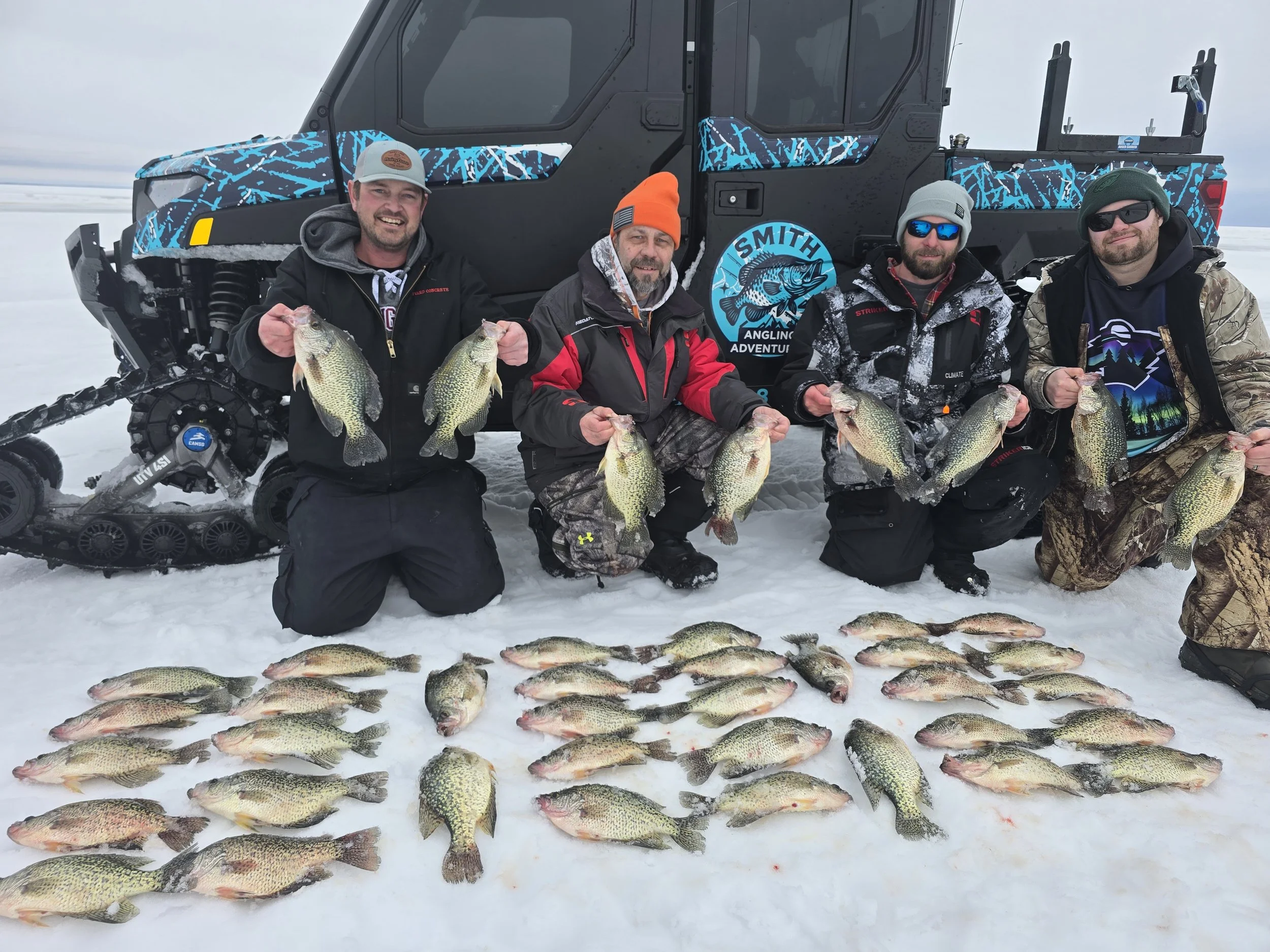 Four men in winter clothing displaying their catch of fish in front of an all-terrain vehicle on snow, with fish laid out on the snow in front of them.