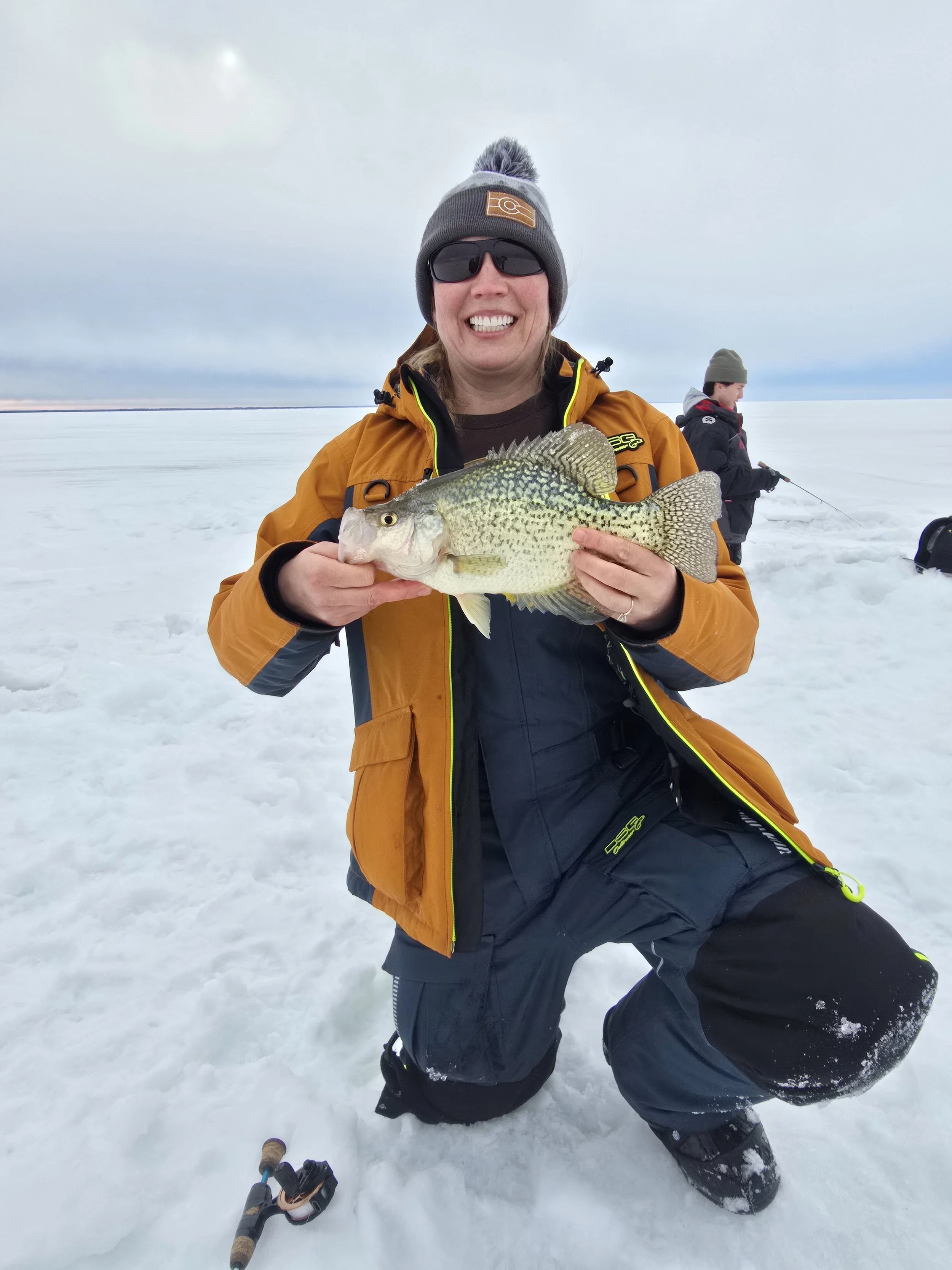 A woman in winter clothing holding a fish in a snowy outdoor setting, with another person fishing in the background.