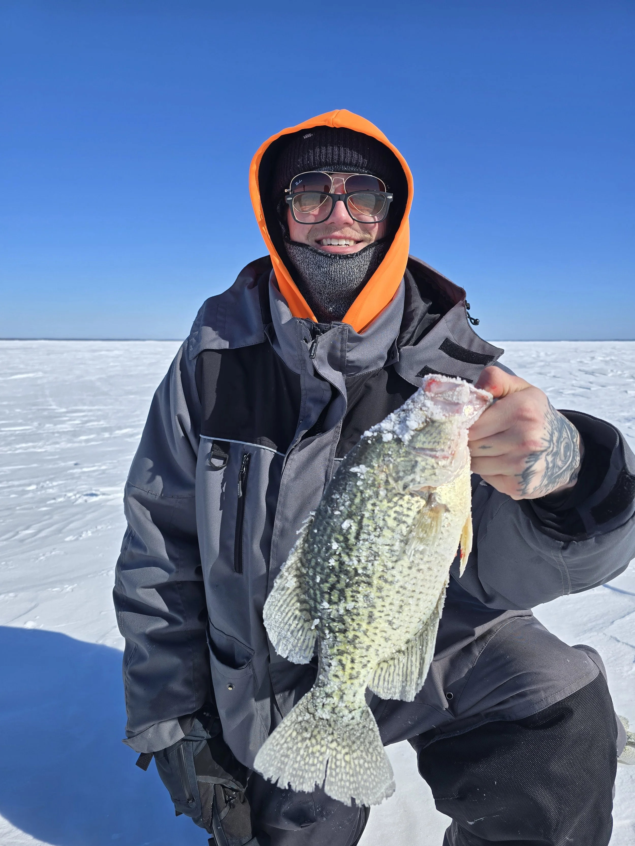 Man in winter gear holding a fish on a snowy landscape with a clear blue sky.