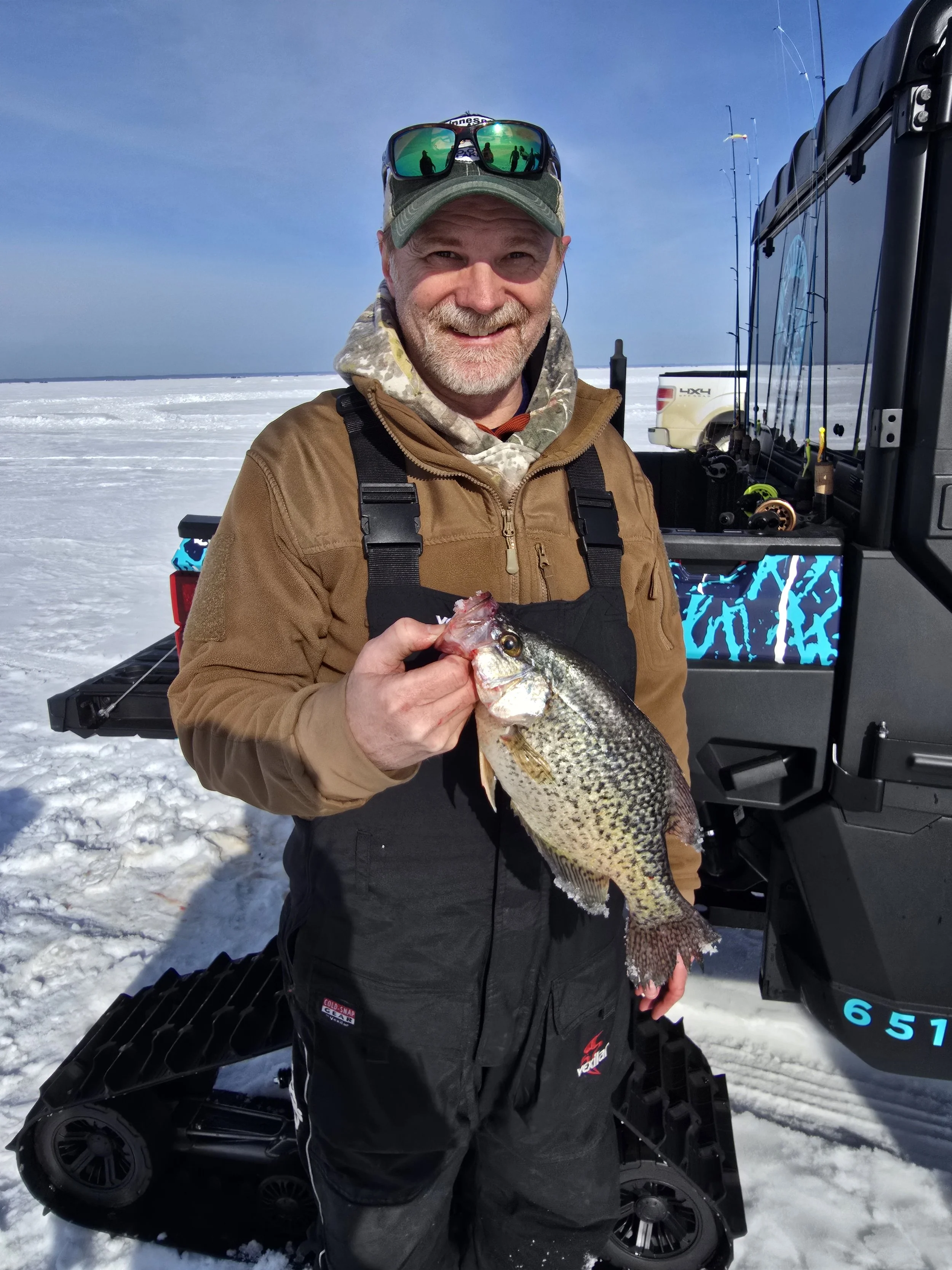 A man in winter clothing holding a large fish on a snowy landscape with fishing rods and vehicles in the background.