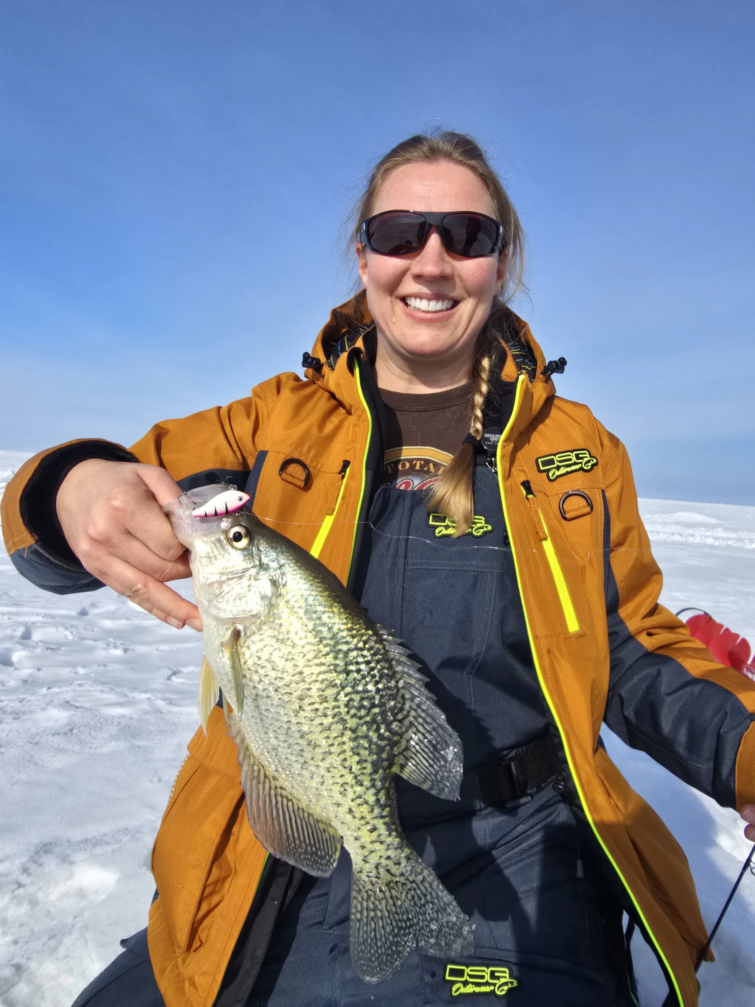 A woman smiling and holding a fish outdoors on snow, wearing sunglasses and an orange jacket.