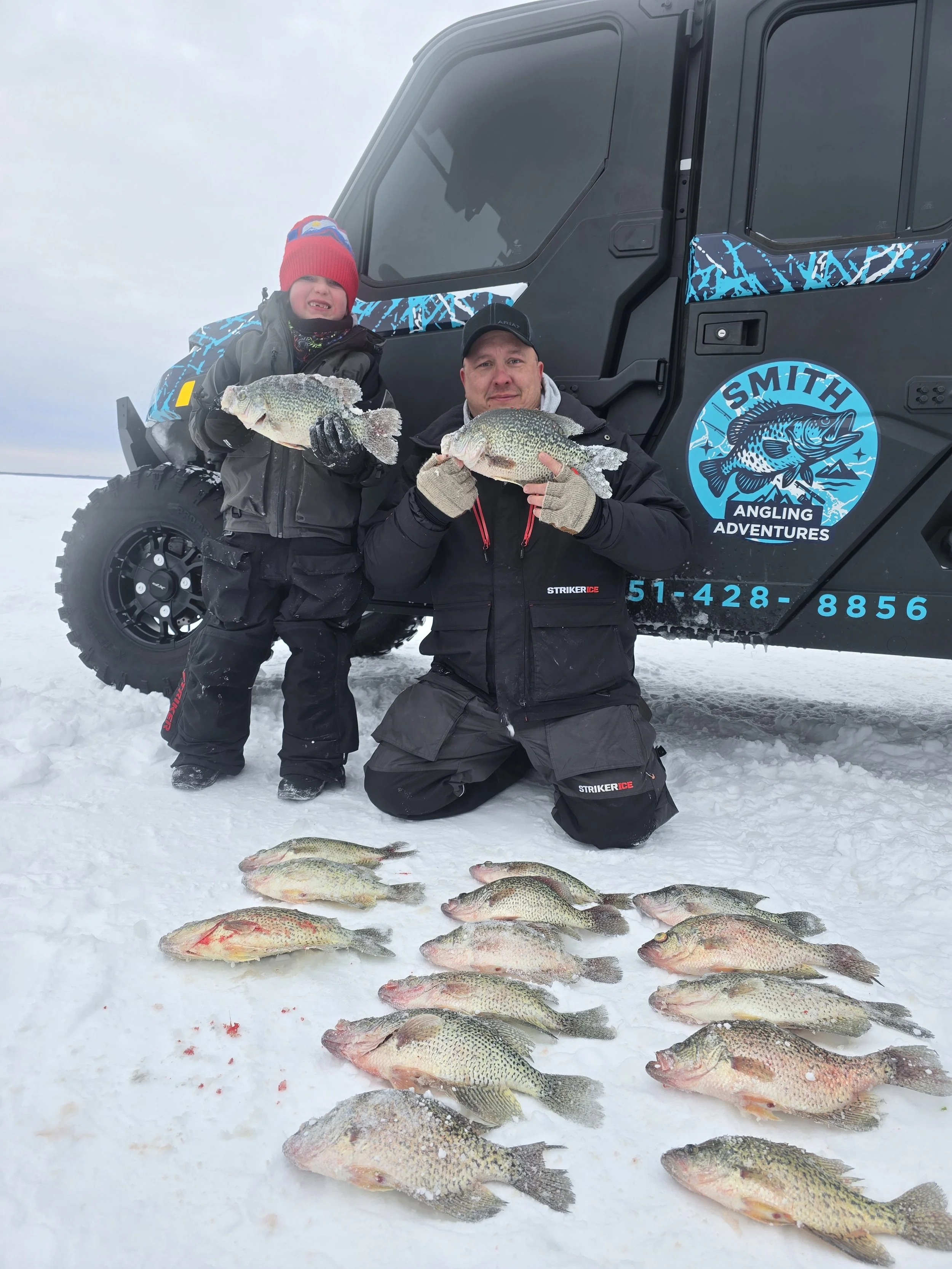 A man and a boy holding fish in front of a black off-road vehicle with a blue logo that says 'Smith Angling Adventures' on a snowy landscape.