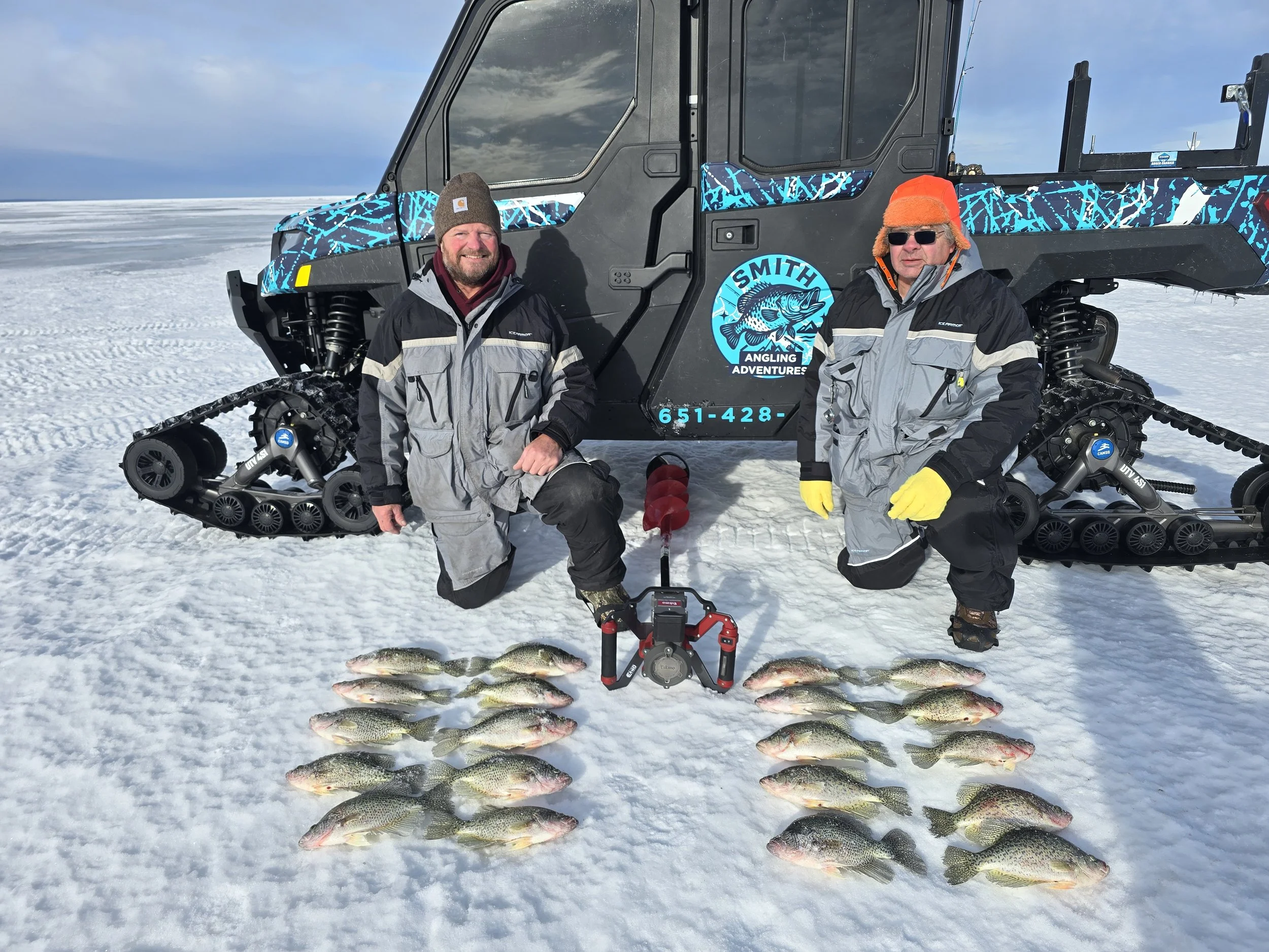 Two men in winter clothing kneeling on snow in front of a black ATV with a blue and black design, featuring the logo 'Smith Angling Adventures,' and a phone number. Several caught fish are laid out on the snow in front of them.