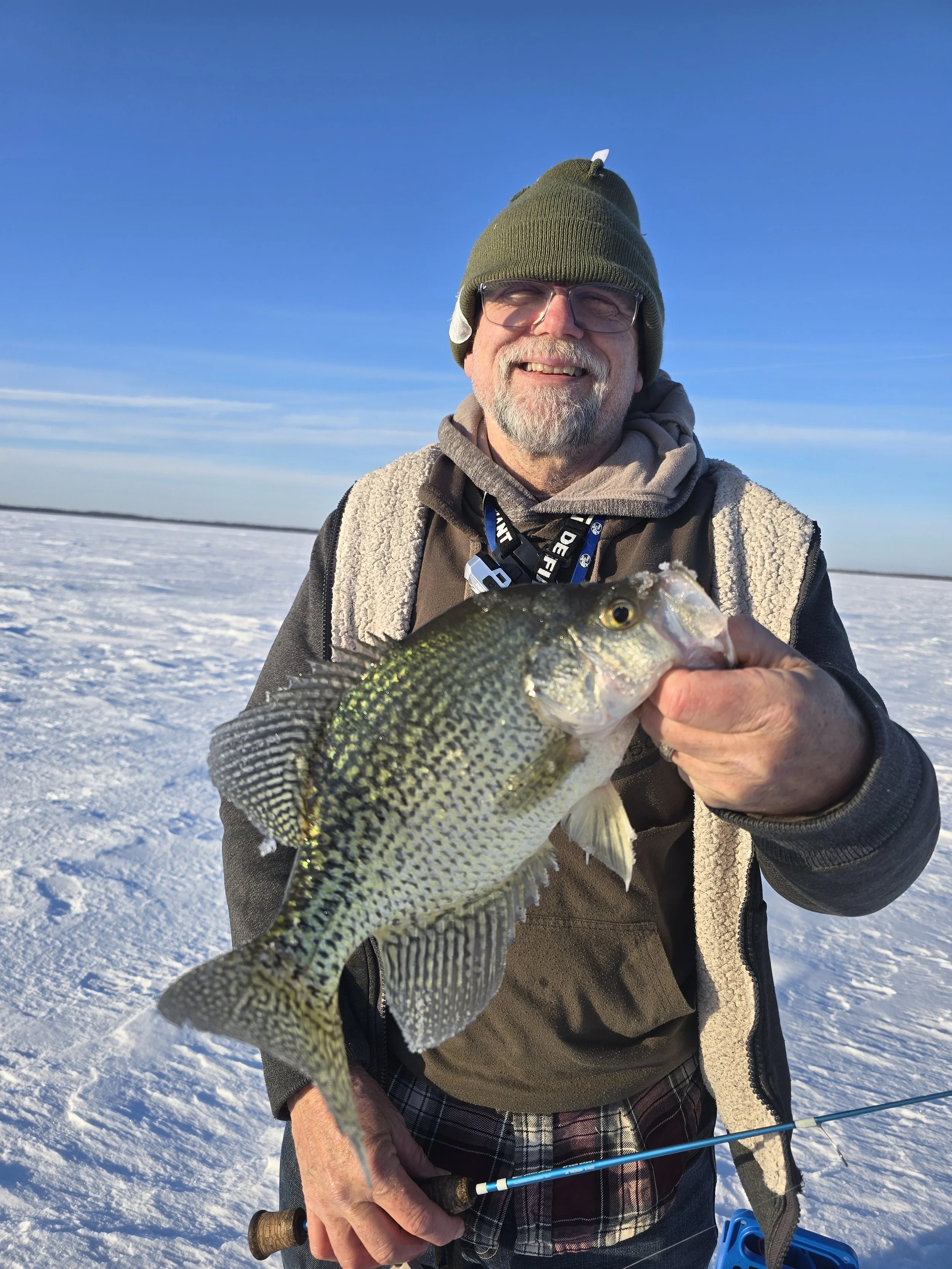 An elderly man with glasses, a green beanie hat, and a warm jacket, smiling while holding a fish on a snowy landscape during daytime.