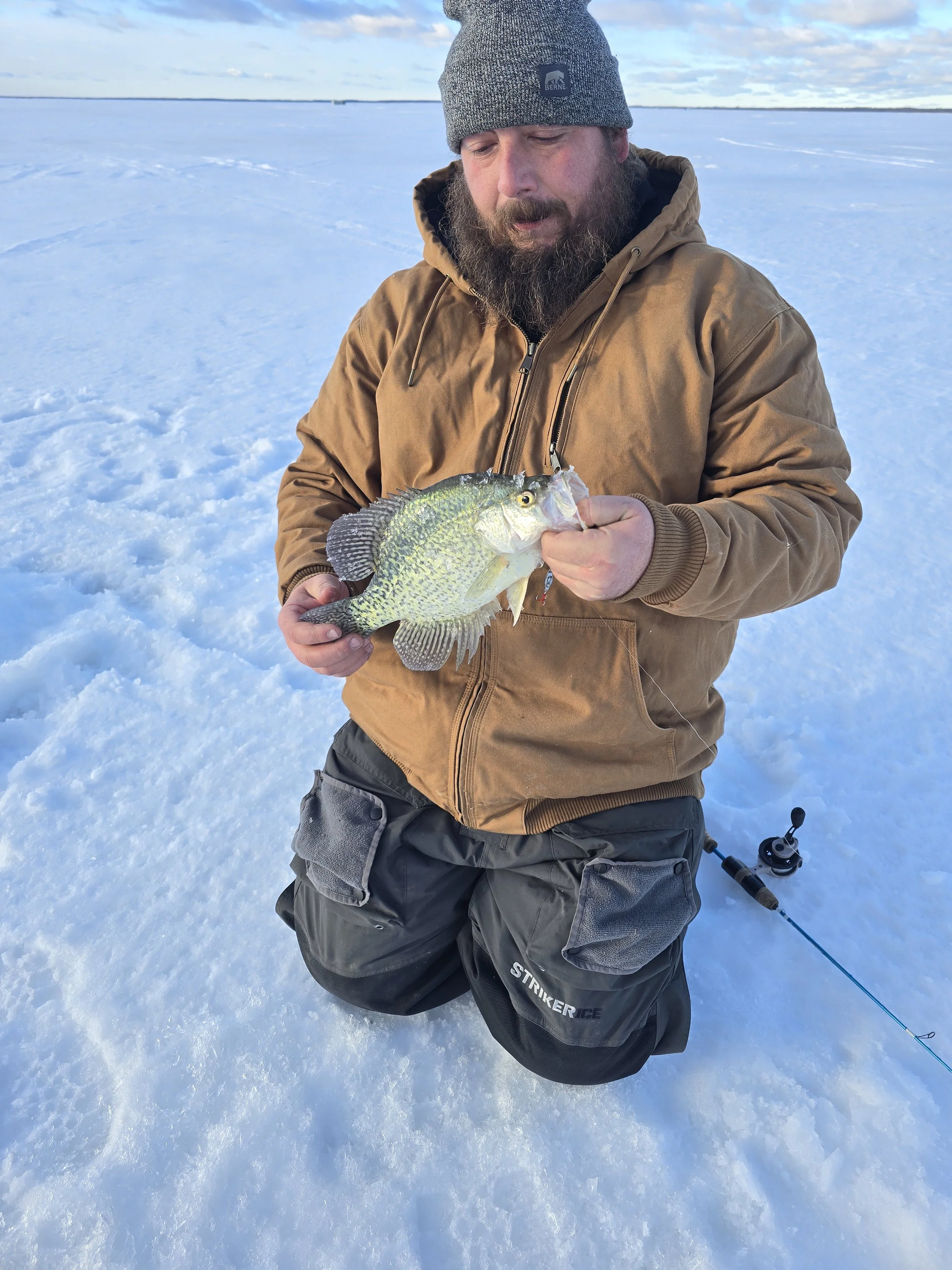 A man kneeling on snow holding a fish with a fishing rod nearby, wearing a brown jacket and gray beanie.