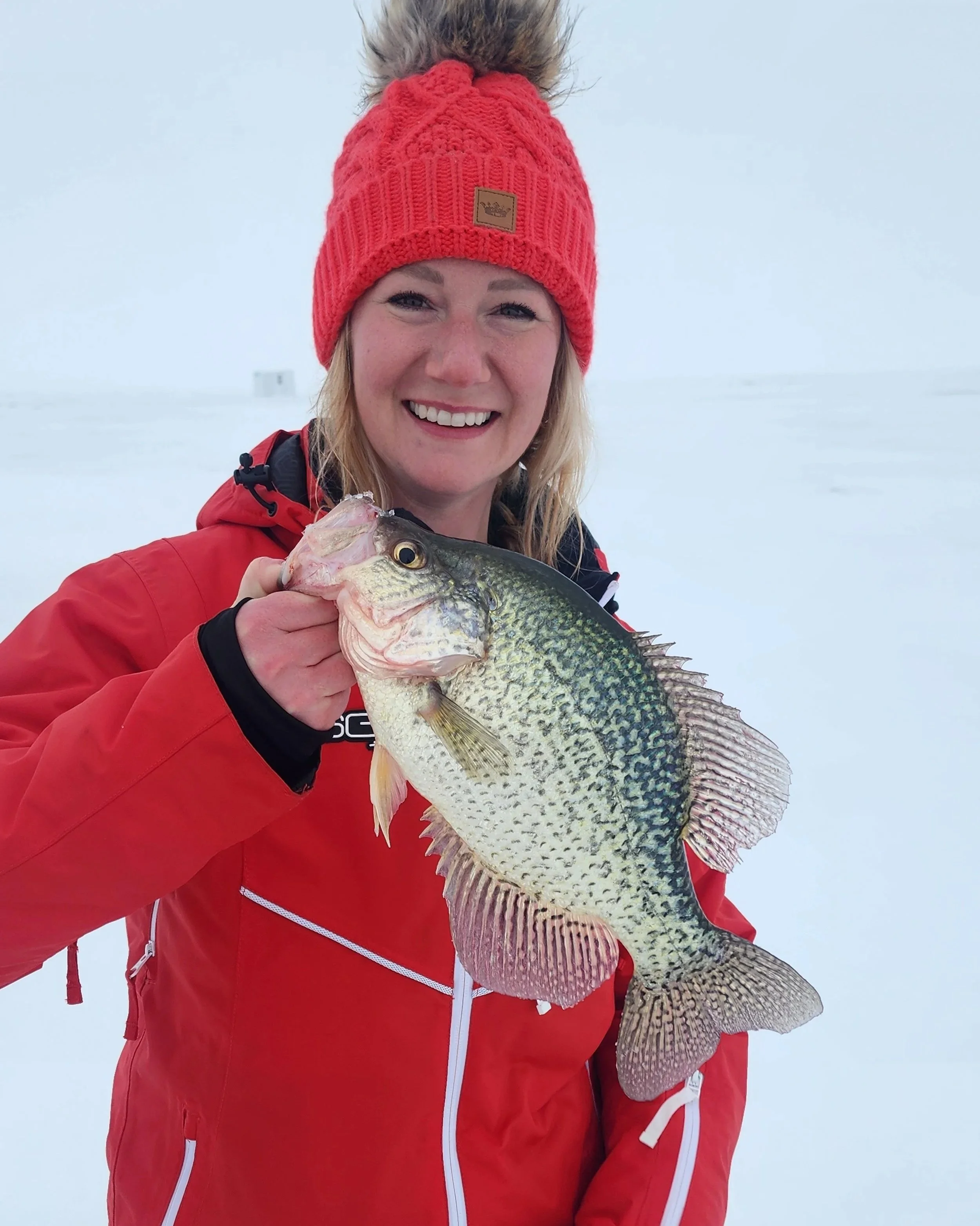 Person wearing a red knit hat with a pom-pom on top and a red jacket, holding a large fish outdoors in a snowy environment