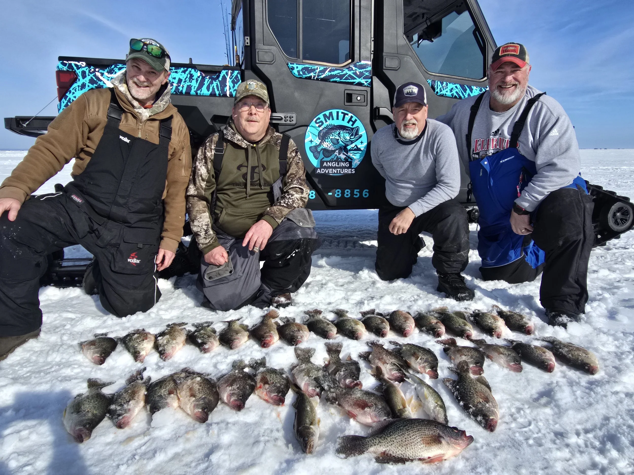 Five men in outdoor winter gear kneeling and standing on snow next to a large catch of fish. Behind them is a black and blue all-terrain vehicle with a logo for 'Smith Angling Adventures' on the side.
