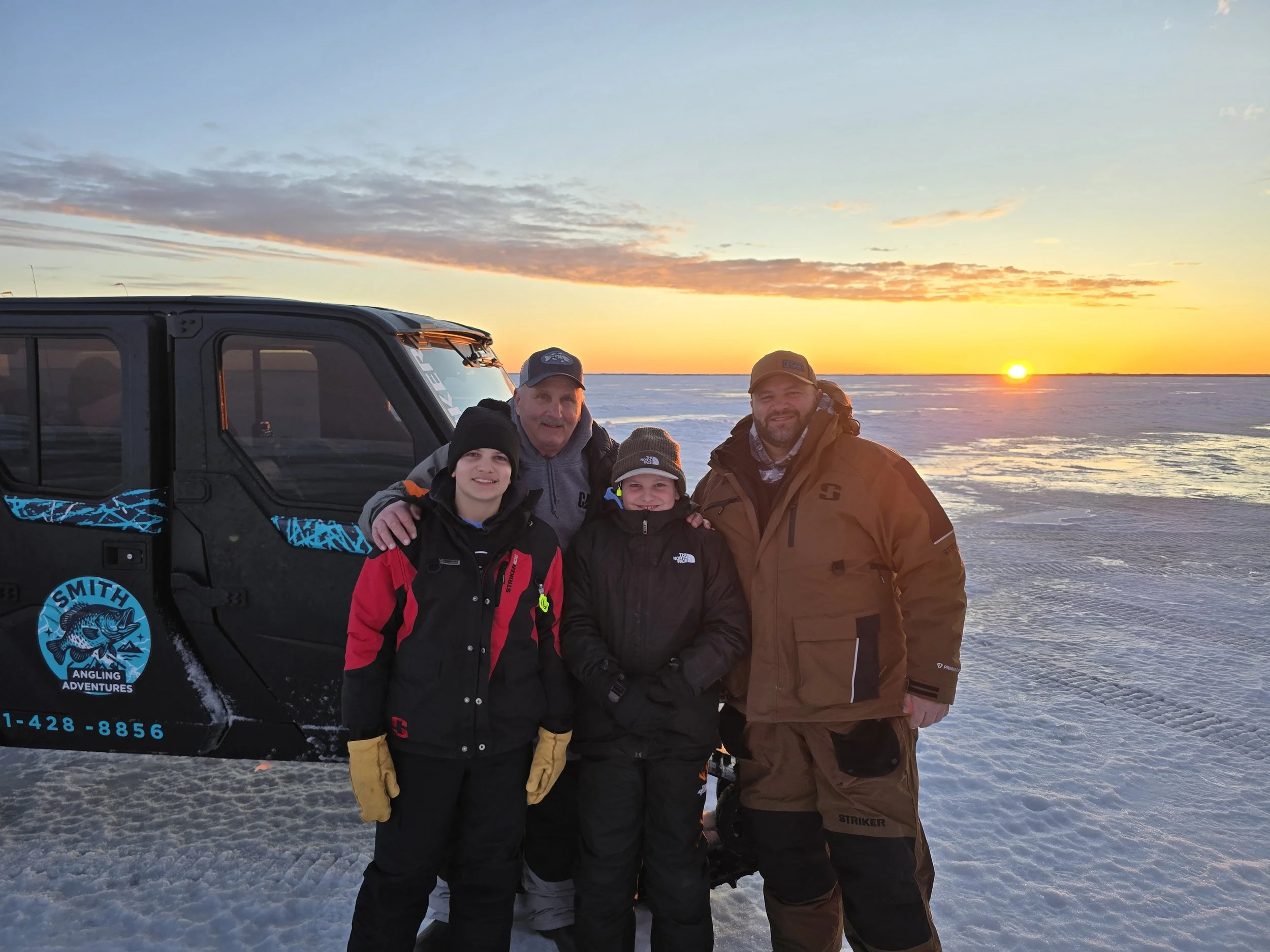 Four people standing together outdoors during sunset in a snowy landscape, with a black off-road vehicle nearby, wearing winter clothing and smiling.