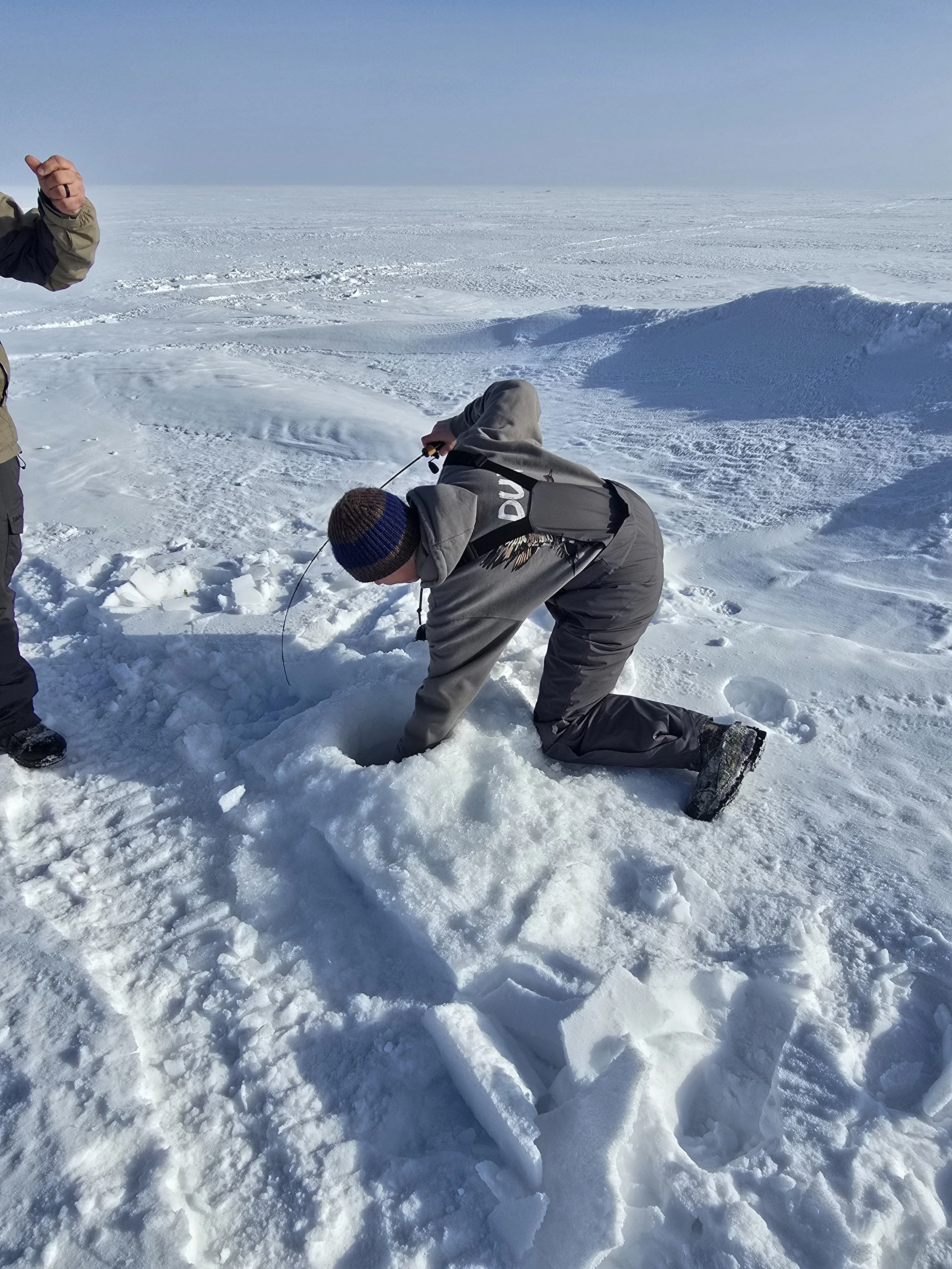 A person kneeling in snow while holding a small container or object, with another person standing nearby in a snowy landscape under a clear sky.