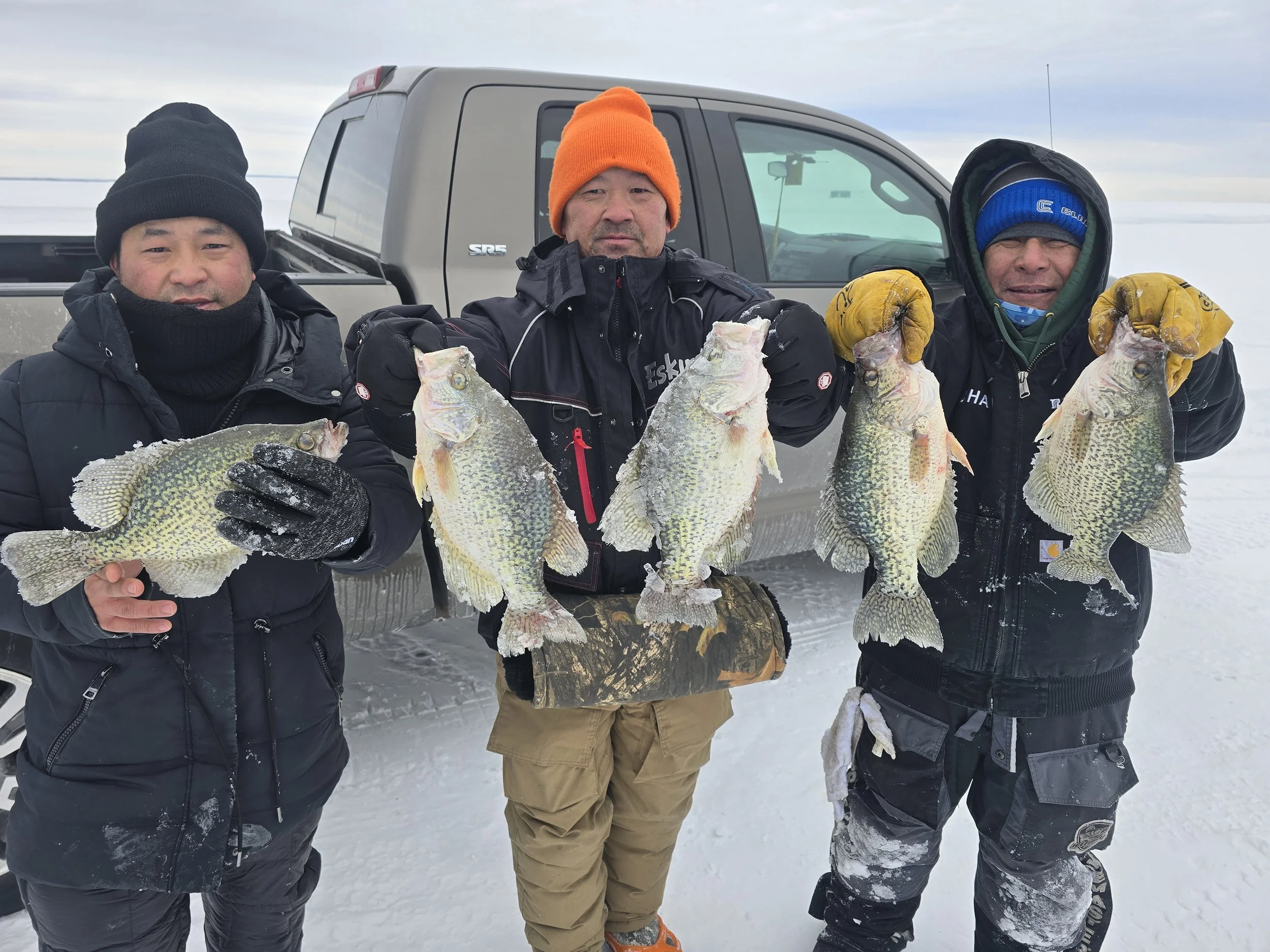 Three men in winter clothing holding large fish on a snowy landscape with a truck in the background.
